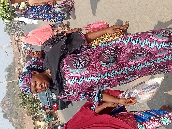Several women wearing brightly patterned dresses and head coverings are lying on a paved surface, possibly a road. One woman holds a sack labeled "Dangote Cement". A black vehicle is partially visible in the background along with buildings and a hill in the distance under a clear sky. The scene appears to be outdoors in a busy area.
