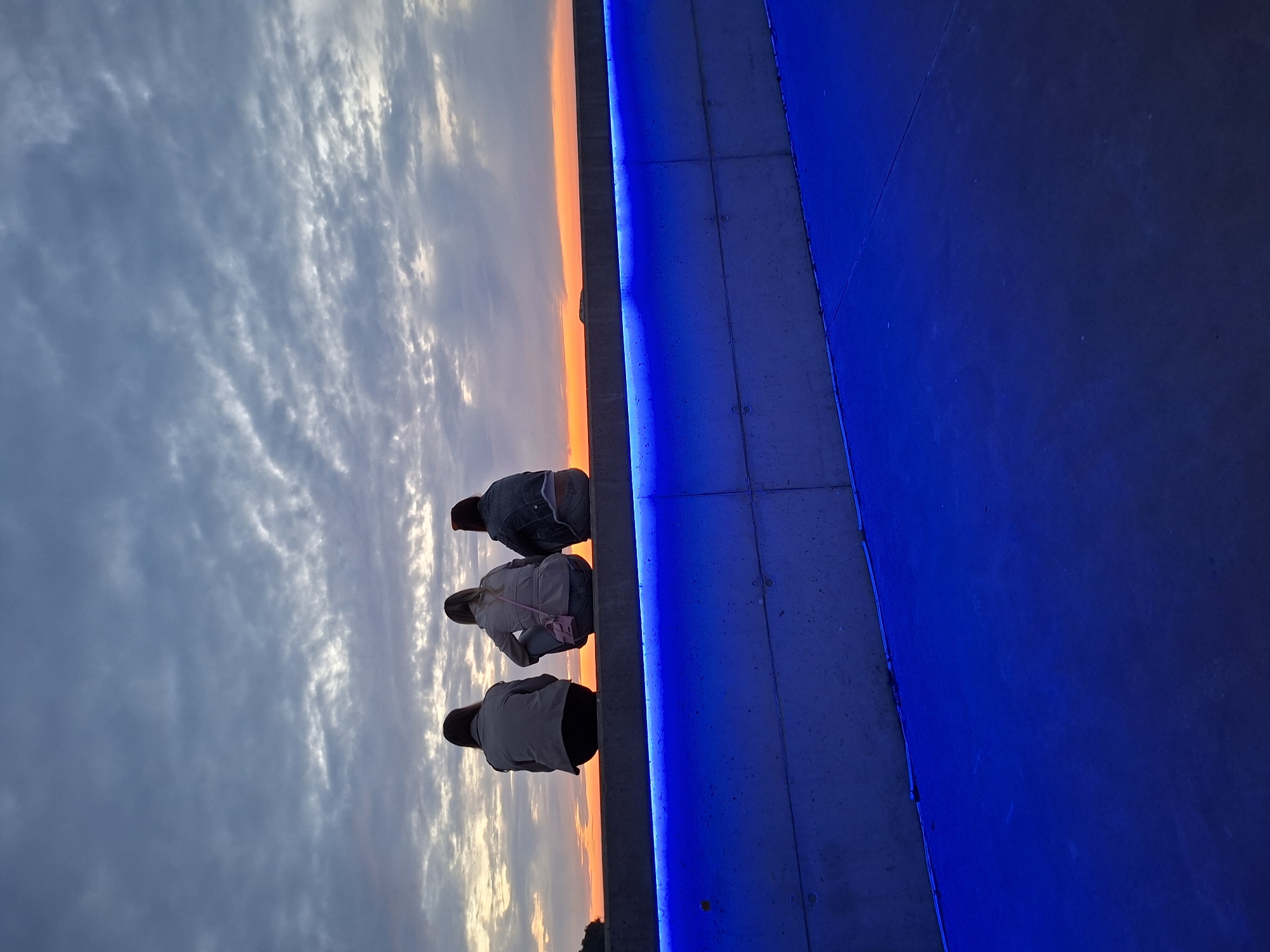The image displays an atmospheric outdoor scene, likely at sunset or sunrise, featuring three individuals observing a vibrant horizon. The left side of the frame is dominated by a dramatic sky, characterized by textured grey clouds above and a brilliant, fiery orange and yellow glow along the horizon line.
Three people, seen from behind, are crouched or sitting on a low ledge, their backs to the viewer. They are focused on the captivating view of the sky. Their attire is casual, with one person wearing a denim jacket and another accessorized with a small pink crossbody bag. Their posture conveys a shared moment of calm observation.
A distinctive feature of the scene is a prominent, bright blue LED light strip that runs along the edge of the ledge where the individuals are positioned. This artificial blue illumination intensely lights up the adjacent concrete surface, which occupies the right portion of the image. The strong blue light creates a striking visual contrast with the natural warm hues of the sky, blending modern design with a natural spectacle.
The composition highlights the juxtaposition of natural light and man-made illumination. Given the location context of Tallinn, Estonia, this setting could be a contemporary waterfront promenade, a public viewing platform, or an urban area designed for evening ambiance, offering scenic views over the city or water. The image evokes a sense of tranquility and appreciation for the beautiful transition of daylight. There is no visible text in the image.