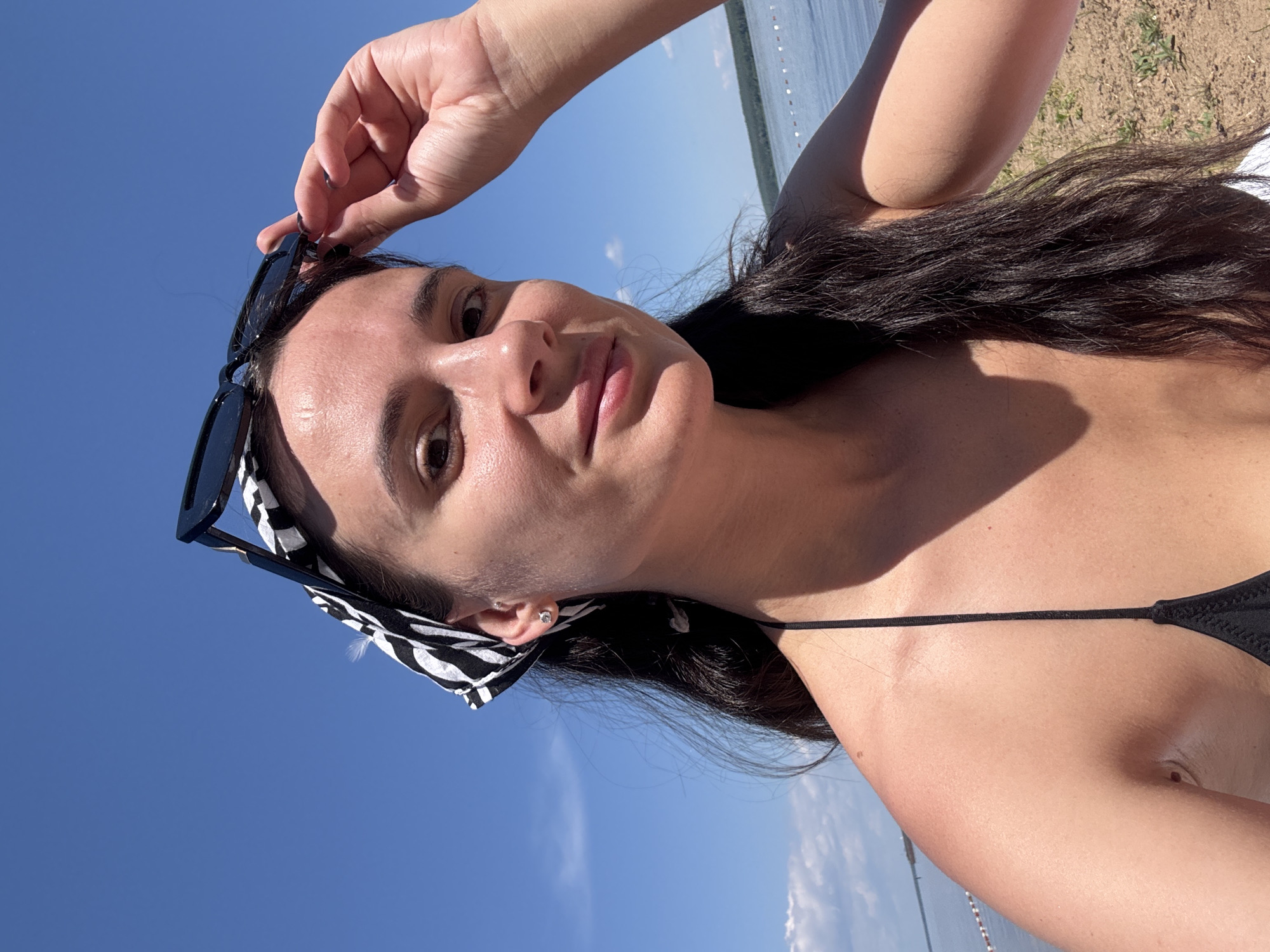 A young woman with long, dark hair, wearing a black bikini top and a black and white patterned bandana, poses for a selfie on a beach in Chaikovsky, Russia. She is holding a pair of black sunglasses up to her face with her right hand. The background shows a bright blue sky with some clouds, a body of water, and a sandy beach with some greenery.