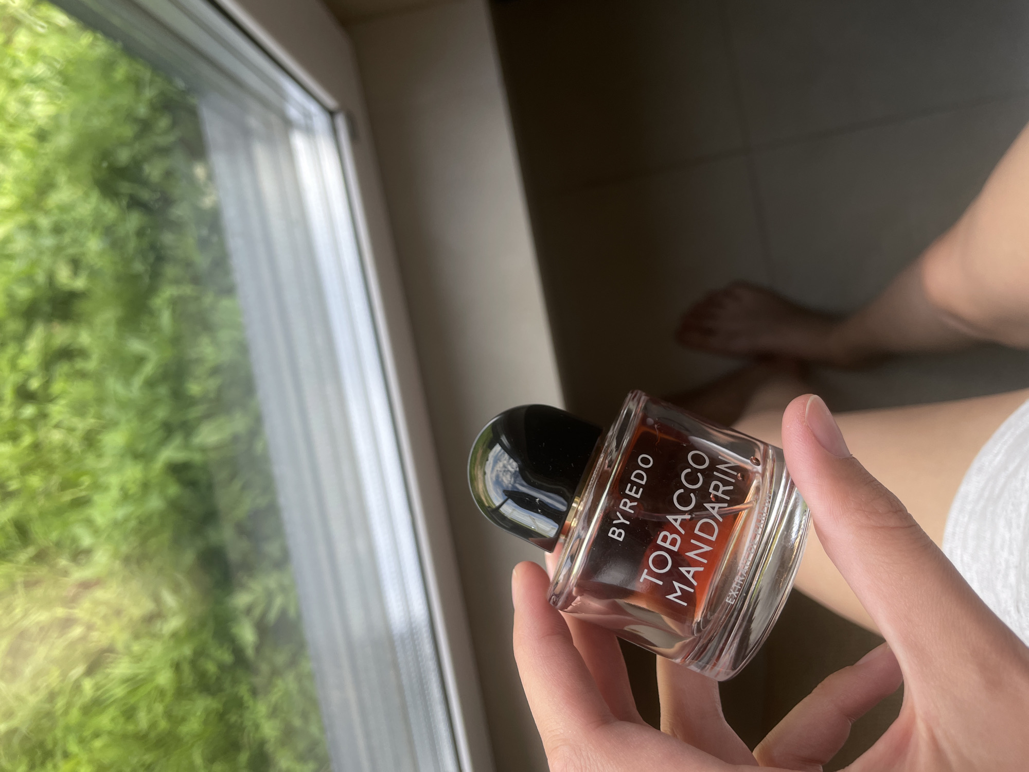 In the city of Podshivalovo, Russia, a hand is holding a clear glass bottle of BYREDO Tobacco Mandarin Eau de Parfum. The bottle contains a reddish-orange liquid and has a black cap. The background shows a window with green foliage visible outside, suggesting an outdoor or nature setting. The lighting appears to be natural daylight, casting soft shadows. The overall scene is casual and personal.