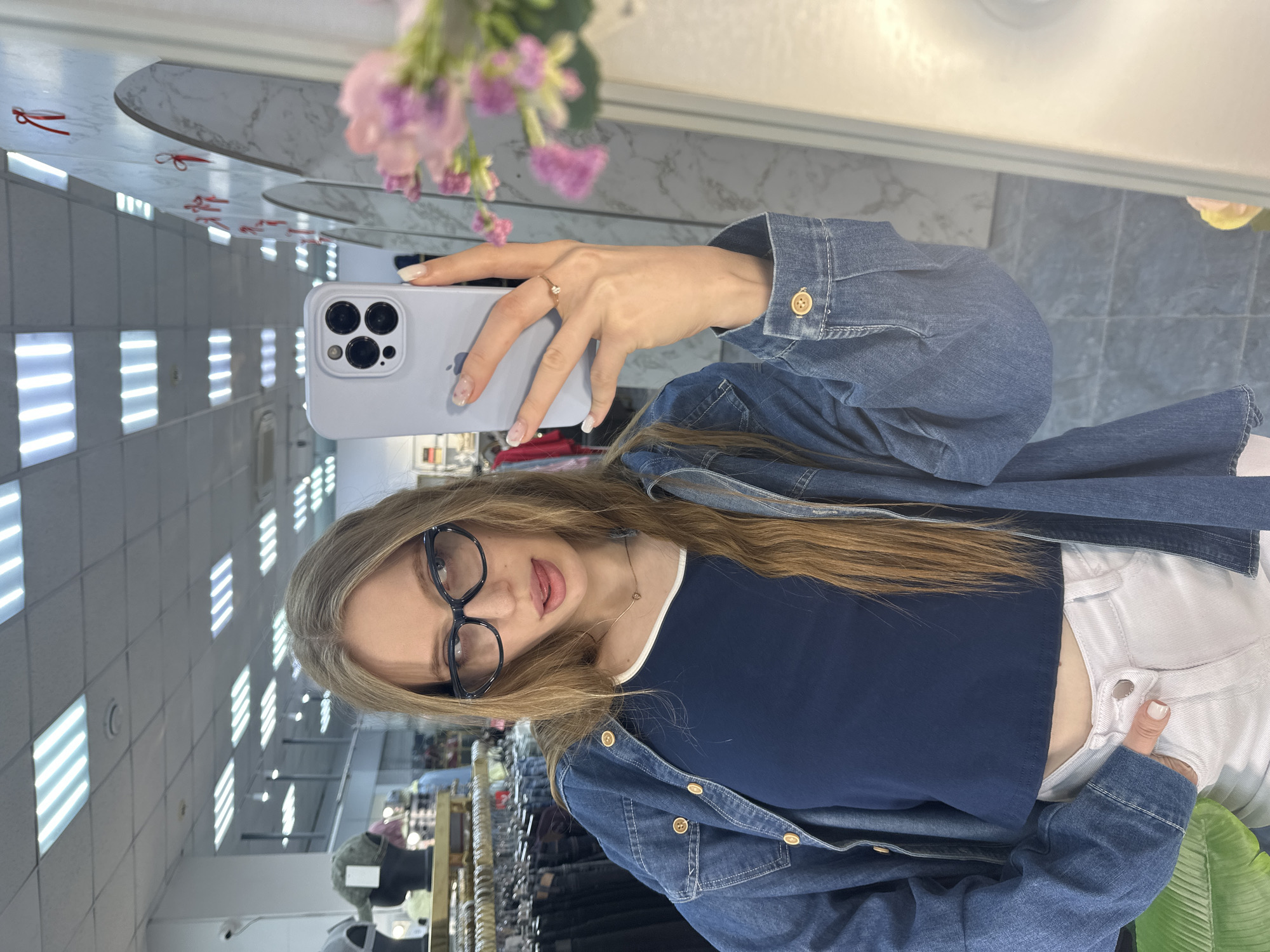 A young woman with long, light brown hair and black-framed glasses is captured in a mirror selfie, holding a light purple iPhone with her right hand. She wears a dark blue crop top, a denim jacket with golden buttons, and light-colored pants. A small heart pendant necklace is visible, and she has a ring on her right ring finger. Her nails are manicured. She has a playful expression with her tongue slightly out.
The selfie reflects the interior of what appears to be a clothing store or boutique in Chaikovsky, Russia. The background shows rows of rectangular fluorescent lights on the ceiling, reflected in the mirror. Above the lights, a light-colored, possibly marble-patterned wall is visible, adorned with small red ribbons. Pink flowers are also reflected, adding a decorative touch. Clothing racks with various garments are visible in the deeper background, along with a small security camera. The floor in the reflection is tiled in a dark grey or blue hue. The overall impression is a casual, indoor setting, likely a commercial establishment.