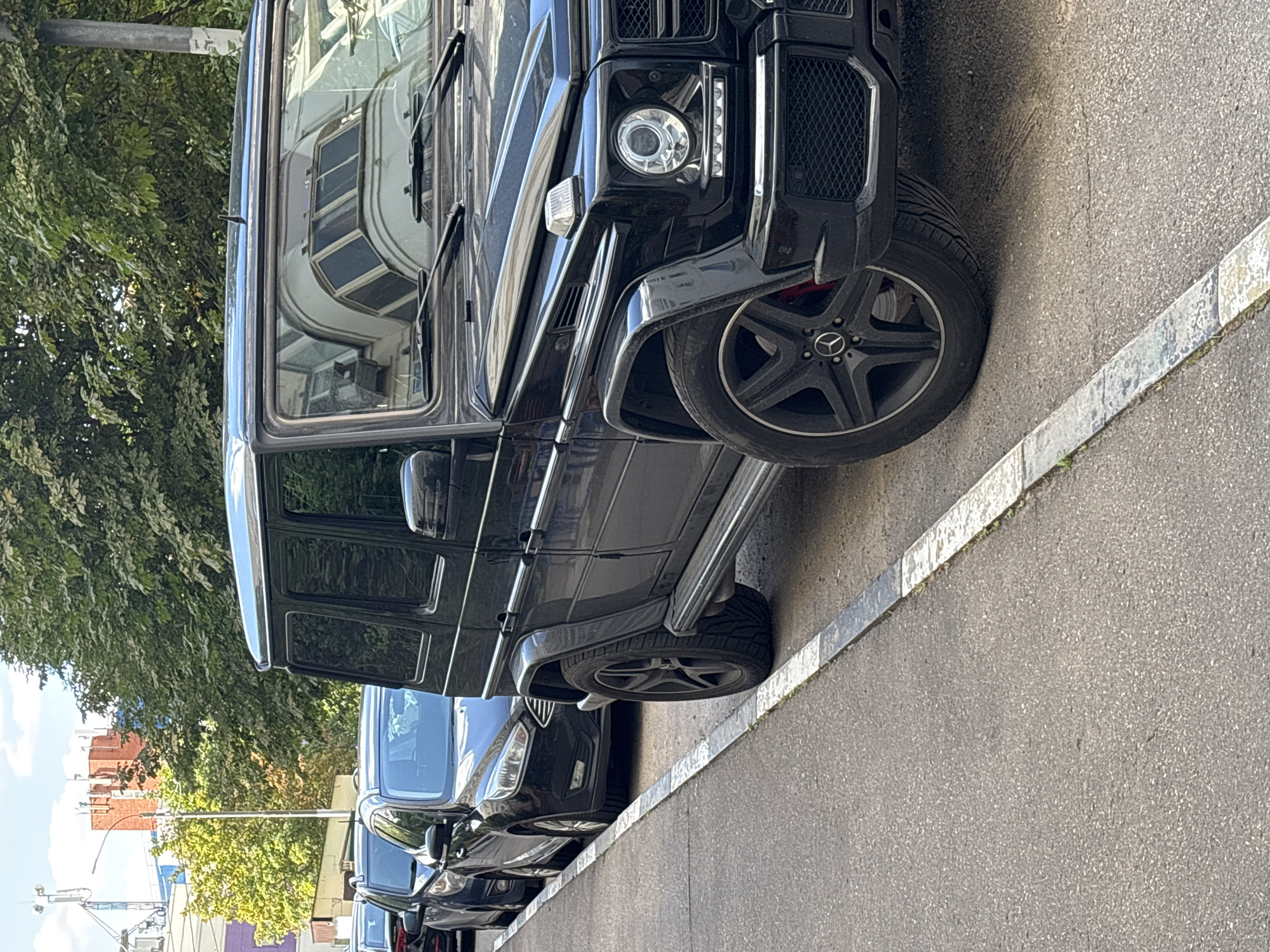 A black Mercedes-Benz G-Class SUV is parked on the side of a city street in Odintsovo, Russia. The SUV is the dominant subject, occupying the foreground and right side of the image. It is angled towards the viewer, showing its front right wheel, fender, and part of its hood and side. The car is parked in a line with other vehicles behind it, including a black Ford Focus. The vehicles are parked on asphalt pavement alongside a curb marked with white and grey stripes. Trees with green foliage line the background, and in the distance, buildings and cell towers are visible. The sky is partly cloudy. There are no people visible in the image.