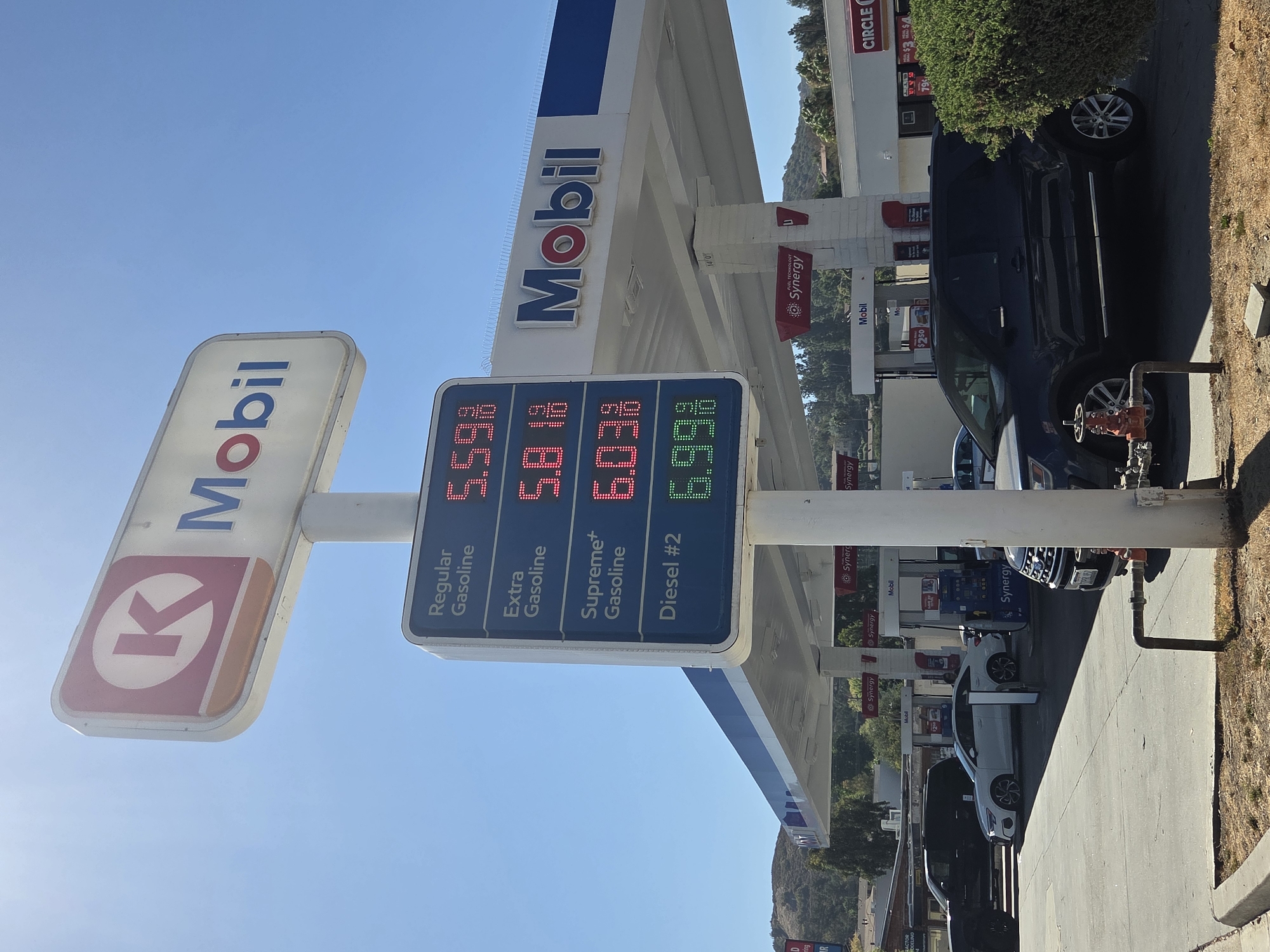 The image depicts a gas station sign displaying fuel prices, with a "K" logo and "Mobil" branding. The sign shows prices for Regular Gasoline at 5.59%, Extra Gasoline at 5.81%, Supreme+ Gasoline at 6.03%, and Diesel #2 at 6.99%. Underneath the sign, several cars are visible, including a white sedan and a dark SUV. The gas station canopy is also visible, with more "Mobil" and "Synergy" branding. The background shows a hilly landscape with trees and buildings under a clear blue sky. The scene appears to be in an outdoor, daytime setting. The image is taken from a ground-level perspective.
