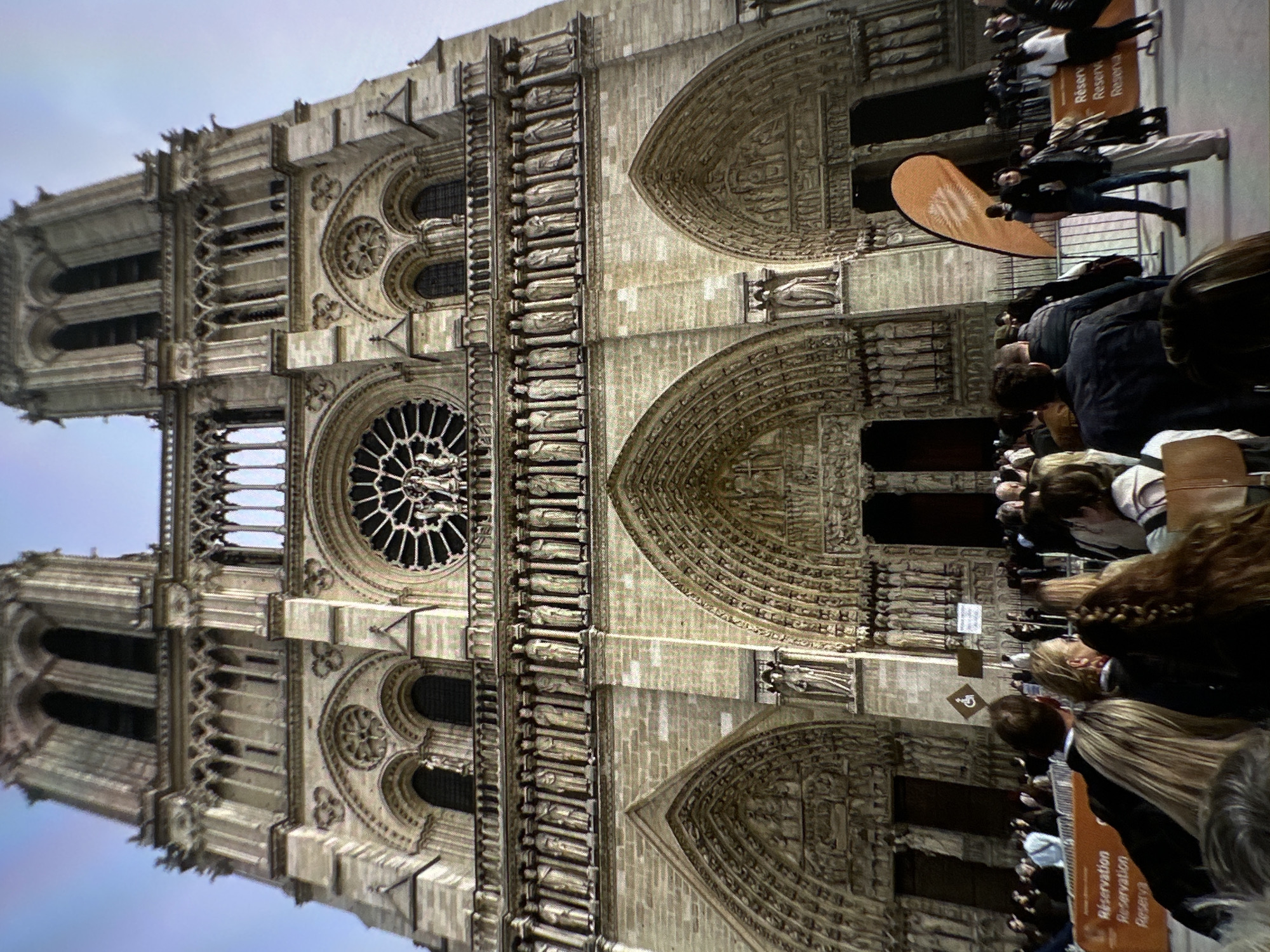 The image shows the façade of Notre Dame Cathedral in Paris, France. The Gothic architecture is visible, including the rose window, towers, and arched portals. A crowd of people is standing in front of the cathedral, with reservation and registration signage displayed. There is no indication that this image is related to Chișinău, Moldova.