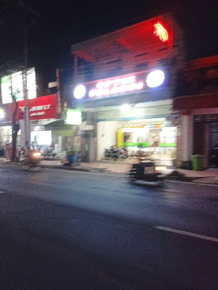 The image depicts a blurry, low-light scene of a street lined with commercial buildings in Mojosari, Indonesia, at night. Artificial lights from various shop signs illuminate the immediate foreground and buildings, contrasting with the dark sky.
On the left, a building features a red sign with partially visible white text. The upper part of the text clearly reads "FLY," and below it, "Auto Shop" or "Audo Shop" is faintly discernible. Above this, a rectangular sign glows green.
In the center-right, a prominent illuminated sign horizontally spans a shopfront. The upper part of this sign clearly displays "OPTIK," indicating an optical store. Below "OPTIK," the word "PELANGI" is partially visible. Above this main sign, a red neon sign, possibly a stylized hand or a brand logo, is brightly lit.
Several motorcycles or scooters are parked on the sidewalks in front of the shops. Another vehicle, possibly a scooter, appears to be on the asphalt road in the foreground, with motion blur indicating movement or recent stopping. Two green trash bins are visible near the right side of the frame. The road surface appears dark and possibly damp, reflecting some of the light. No specific individuals are clearly identifiable due to the motion blur and low light conditions. The scene suggests a typical evening in a commercial district.