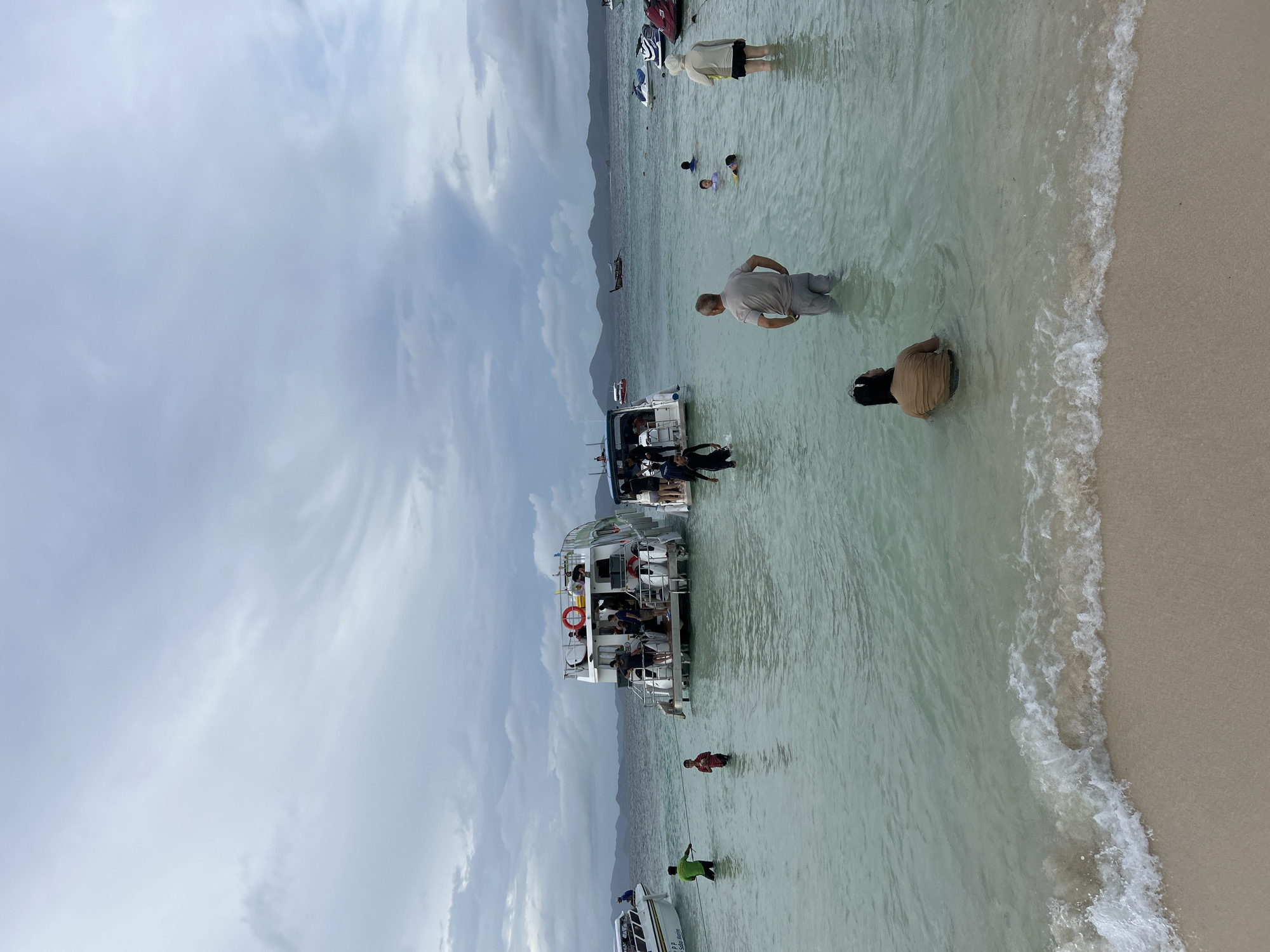 The image captures a beach scene with several boats docked in shallow, clear turquoise water under an overcast sky. In the foreground, a gentle wave breaks on a sandy shore. Several people are visible in the water, wading or swimming. Two white boats with multiple decks are anchored closer to the shore, with some individuals standing on their decks and others in the water around them. Further out, more boats, including a jet ski, are present in the distance. The horizon shows a line of hills or mountains. The overall impression is a popular tourist destination with recreational activities taking place. This scene is consistent with the coastal areas of Tambon Phru Nai, Thailand, known for its beaches and island hopping tours.