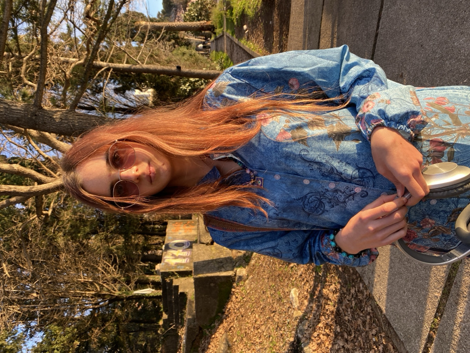 The image, rotated 90 degrees clockwise, depicts a young person outdoors in what appears to be a park or natural setting, bathed in warm, golden sunlight. The person, positioned slightly to the left of the frame, has long, flowing reddish-brown hair that cascades over their shoulders and chest. They are wearing round, pink-tinted sunglasses, which obscure part of their eyes but show a subtle smile on their face.
The individual is dressed in a long-sleeved, collared garment, possibly a dress or a long shirt, made of a light blue fabric. The fabric features intricate, ornate patterns in darker blues, grays, and pinks, resembling floral motifs and swirls. The sleeves are slightly puffy at the shoulders and cuffed at the wrists. On their left wrist, a multi-beaded bracelet with turquoise-like and darker beads is visible.
Their hands are positioned in front of them, holding a pair of silver and black over-ear headphones. Their right hand is holding one earcup, while their left hand is near the headphone jack or cable.
The background consists of several trees with bare branches, suggesting either late autumn, winter, or early spring. The ground is covered with fallen leaves and some sparse vegetation. To the right and behind the person, there's a concrete or stone structure, possibly a low wall or steps, with some unidentifiable graffiti visible on its surface. The overall lighting indicates a sunny day, likely during the late afternoon or early morning, with warm light illuminating the scene. No discernible text is clearly readable in the image.