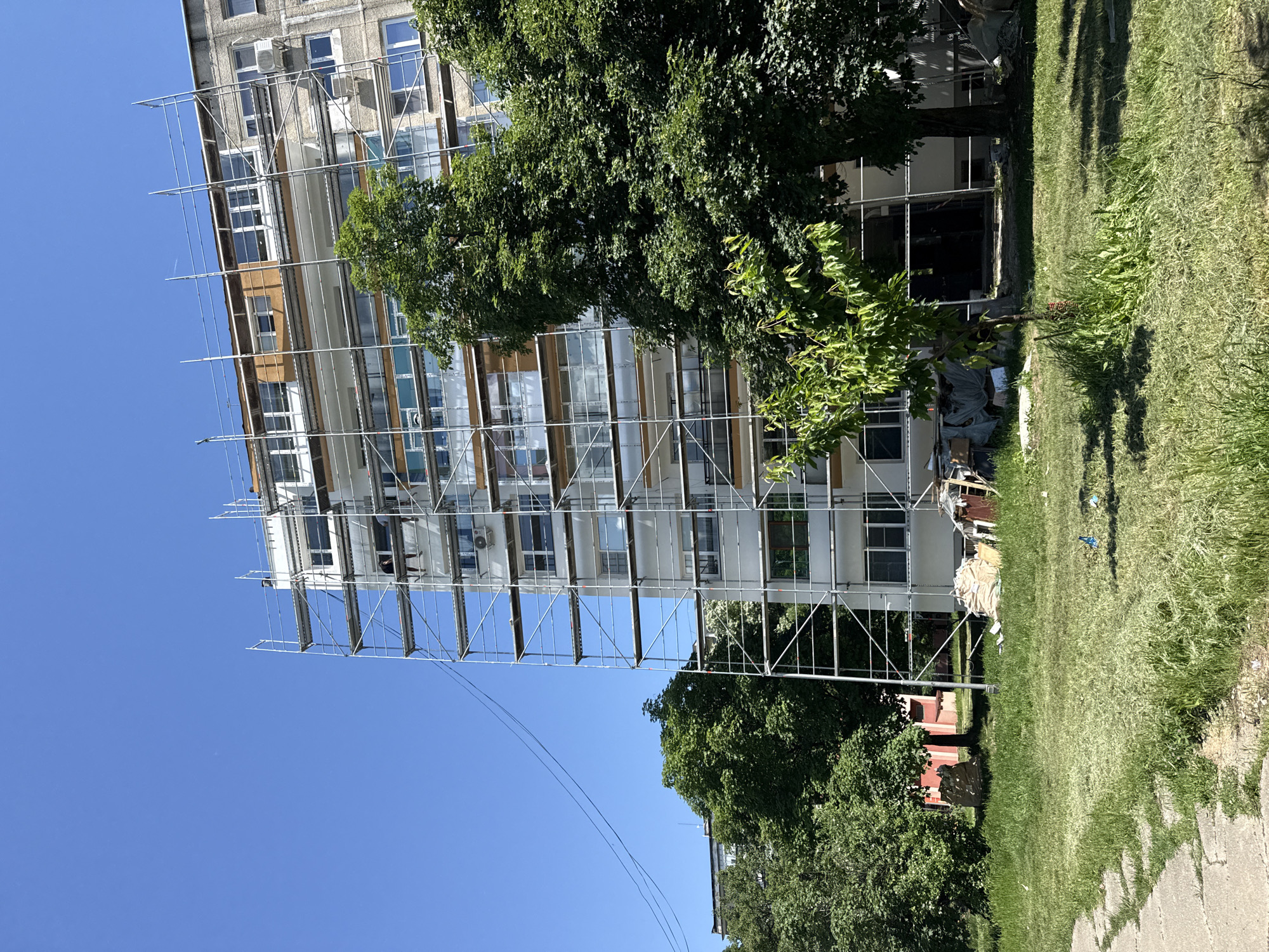 The image depicts a multi-story building in Lovech, Bulgaria, undergoing exterior renovation. Scaffolding covers a large portion of the facade. The building is surrounded by trees and grassy areas. Some construction materials and debris are visible on the ground near the building's base. The sky is clear blue.