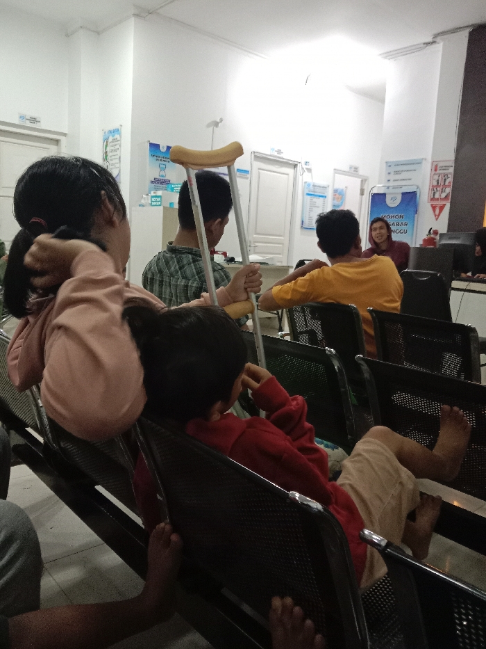 An indoor waiting area in Gladaksari, Indonesia, depicts several individuals seated on black perforated metal chairs. In the immediate foreground, the back of a person with dark hair tied back, wearing a light pink long-sleeved top, is visible; their left arm is raised towards their head. Adjacent, a child in a red long-sleeved top and light-colored shorts sits with bare feet resting on the chair, their head turned away. Further back, a male youth in a plaid shirt holds a silver forearm crutch with a yellow cuff. To his right, another male in a yellow t-shirt is seated, facing away from the camera. In the background, a female staff member or adult in a dark top smiles towards the seated individuals, near a desk with a computer monitor. The white walls are adorned with informational posters in Indonesian, including one stating "MOHON SABAR MENUNGGU" (Please wait patiently). White doors are visible in the background, along with a red fire extinguisher mounted on the wall.