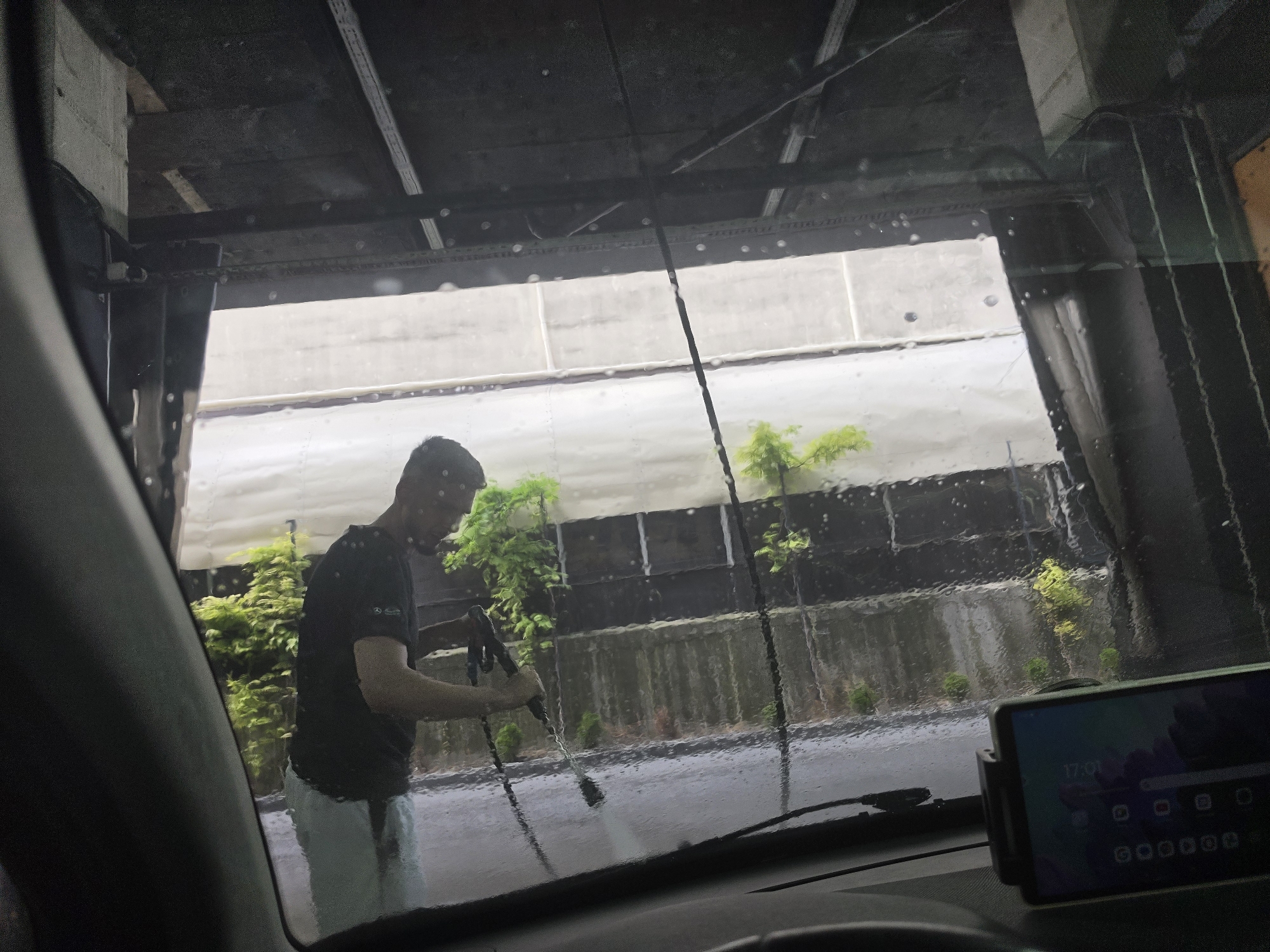 A man is visible from the interior of a vehicle, reflected on the windshield which is covered in water droplets. He is standing outside the vehicle, holding a pressure washer wand and appears to be cleaning something. The background suggests an outdoor car wash area, with lush green trees and shrubs evident, along with a concrete wall. The man is wearing a dark t-shirt and light-colored trousers. A phone mount with a smartphone displaying the time "17:01" is fixed to the dashboard of the vehicle. The scene takes place in Varna, Bulgaria, likely at a self-service car wash.