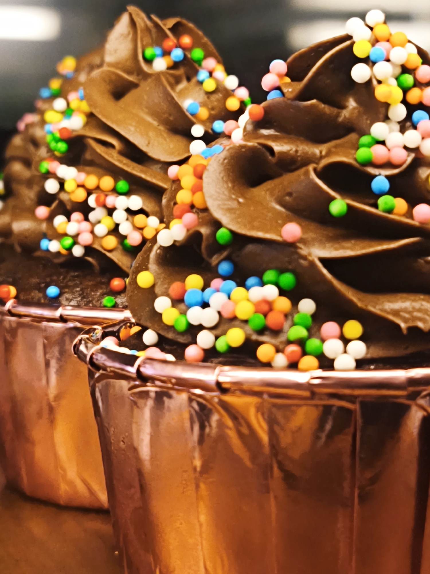 This is a close-up, vertical shot of two chocolate cupcakes with brightly colored sprinkles. The cupcakes are presented in shiny, rose-gold colored liners. The frosting is a rich, dark chocolate, piped into swirled peaks. The sprinkles are small, round, and come in a variety of colors including white, pink, blue, yellow, green, and orange. Some sprinkles are scattered across the frosting, while others are clustered together. The lighting is warm and highlights the glossy texture of the frosting and cupcake liners. The background is softly blurred, suggesting an indoor setting, possibly a bakery or kitchen. No people or text are visible in the image. The focus is entirely on the detailed texture and vibrant colors of the cupcakes, creating an appealing and appetizing presentation.