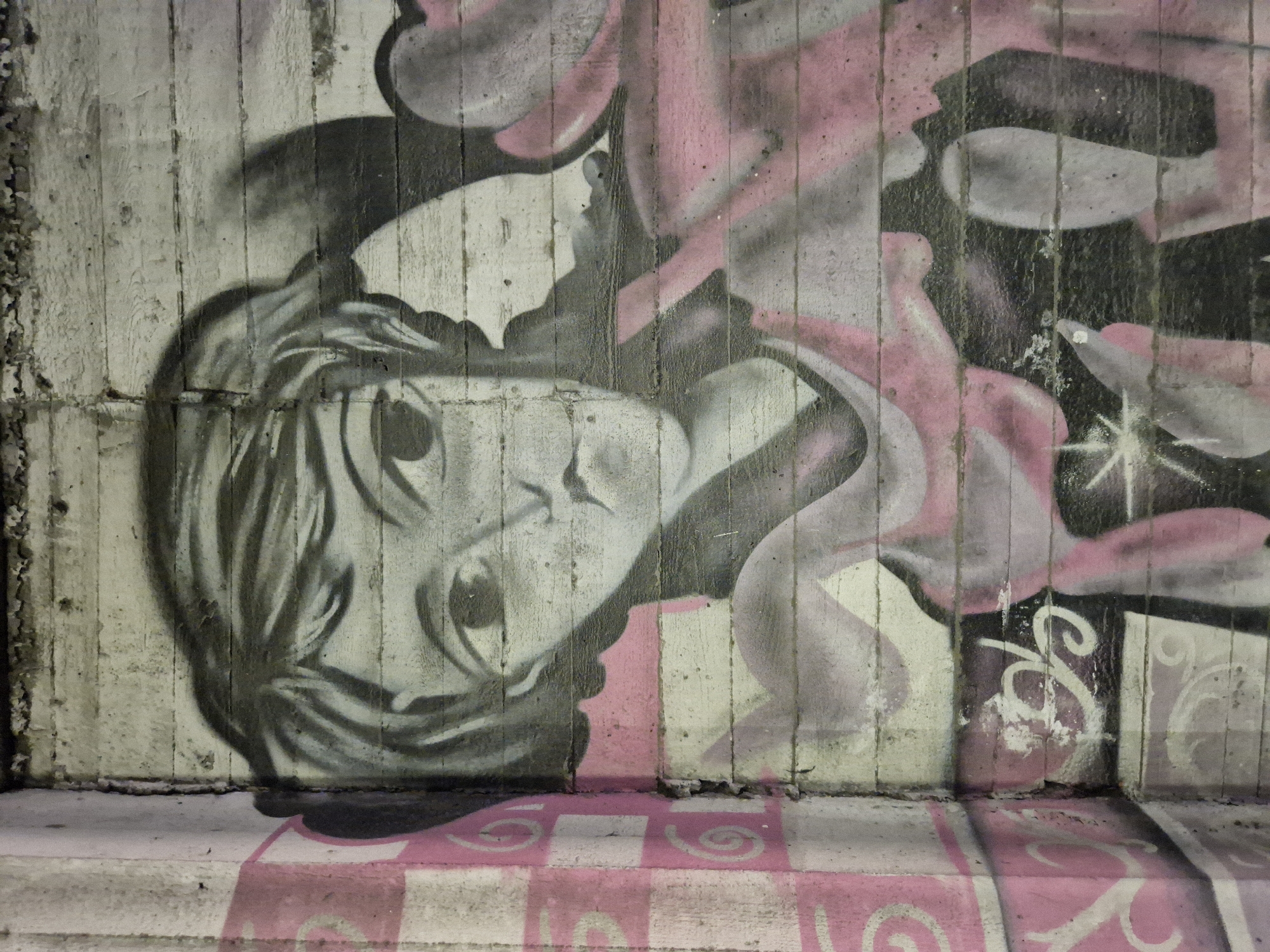 This is a close-up, full-frame, eye-level photograph of a graffiti mural painted on a rough, concrete wall, possibly in the city of Taranto, Italy. The mural depicts a stylized, black and white portrait of a person with large, wide eyes and a slightly melancholic expression. The person's hair is rendered in dark, flowing strokes. Pink accents and shading are incorporated into the mural, particularly around the neck and shoulders, suggesting clothing or a flowing scarf. There is a section of a different graffiti design to the left of the portrait, featuring pink and white checkered patterns with white swirling motifs. The concrete wall itself has horizontal lines, typical of poured concrete construction. The lighting is even, with no strong shadows. The image is sharp and detailed, capturing the texture of the paint and the rough surface of the concrete.