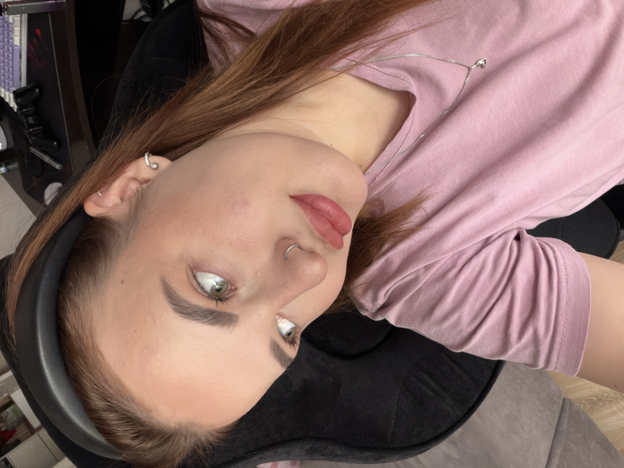 A close-up, diagonally oriented image captures a young woman from the chest up, focusing on her face and upper body in an indoor setting. She has light skin, light brown hair worn down, and striking light green or hazel eyes. Her eyebrows are well-defined, and she wears light makeup, including lip color and eyeliner.
She is adorned with several silver piercings: a helix piercing with a hoop and small stones, a small hoop in her upper earlobe, and a silver septum ring. A delicate silver necklace with a small heart pendant rests on her light pink short-sleeved t-shirt.
The woman is looking upwards and slightly to her left, with a subtle pout on her lips, suggesting a posed expression for the camera. Her head rests against a black, padded surface, possibly a chair back or headrest. The background is partially blurred, revealing hints of household objects like a shelf or desk with items on it to her left, and a light wooden floor to her right. The lighting is soft and even, indicating daytime. There is no visible text in the image. The scene conveys a casual, personal atmosphere, likely a home environment in Krasnodar, Russia.
