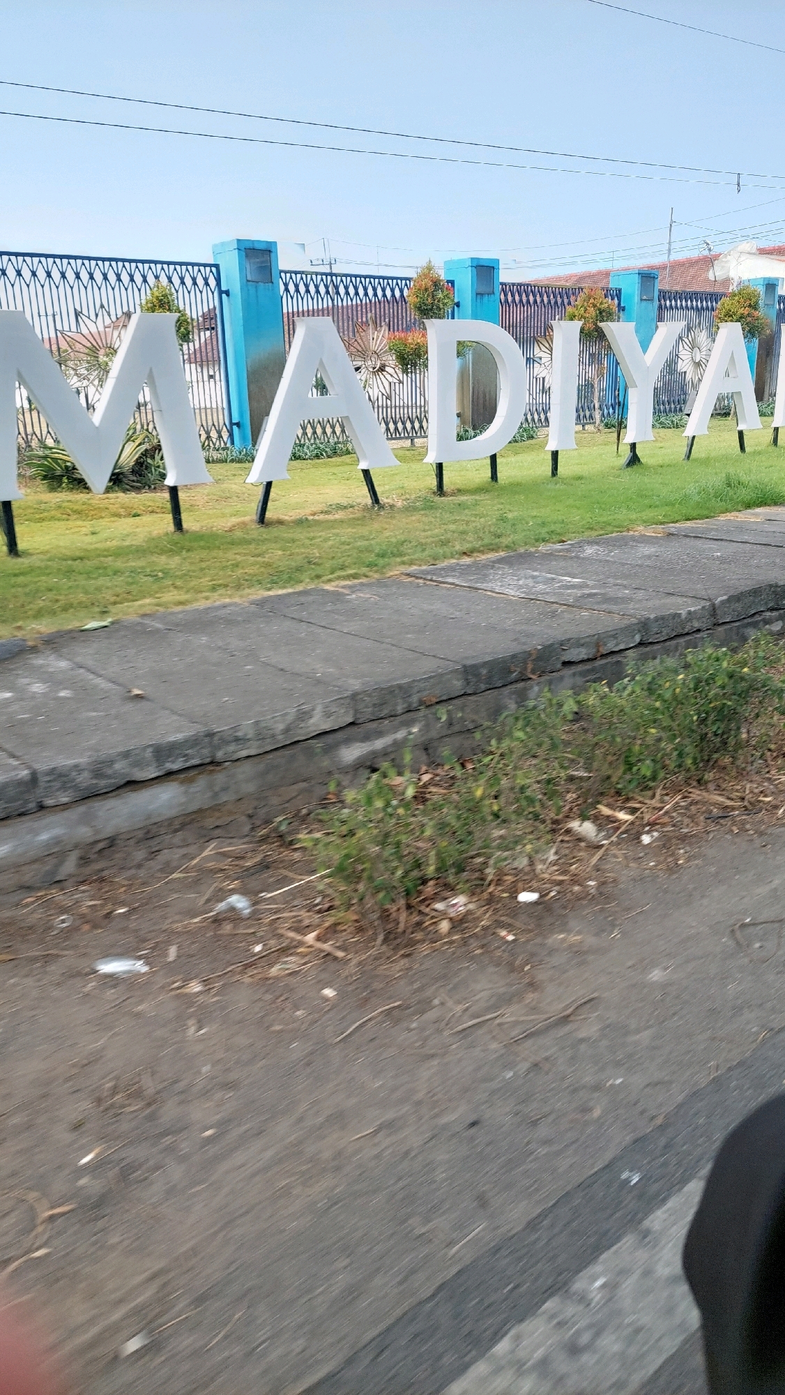 The image shows a close-up view of large, white, three-dimensional letters spelling out "MADIYAR" which are installed in front of a blue fence with ornate metalwork. The letters are standing on individual black poles and are placed on a green lawn. Behind the fence, there are some buildings with red tile roofs and decorative elements. The sky is clear and blue. In the foreground, there is a concrete sidewalk and a paved road, with some dry grass and debris on the side. The overall scene appears to be an entrance or landmark in a public area. It is not possible to determine the specific location in Sidoarjo, Indonesia, from the visual information provided.