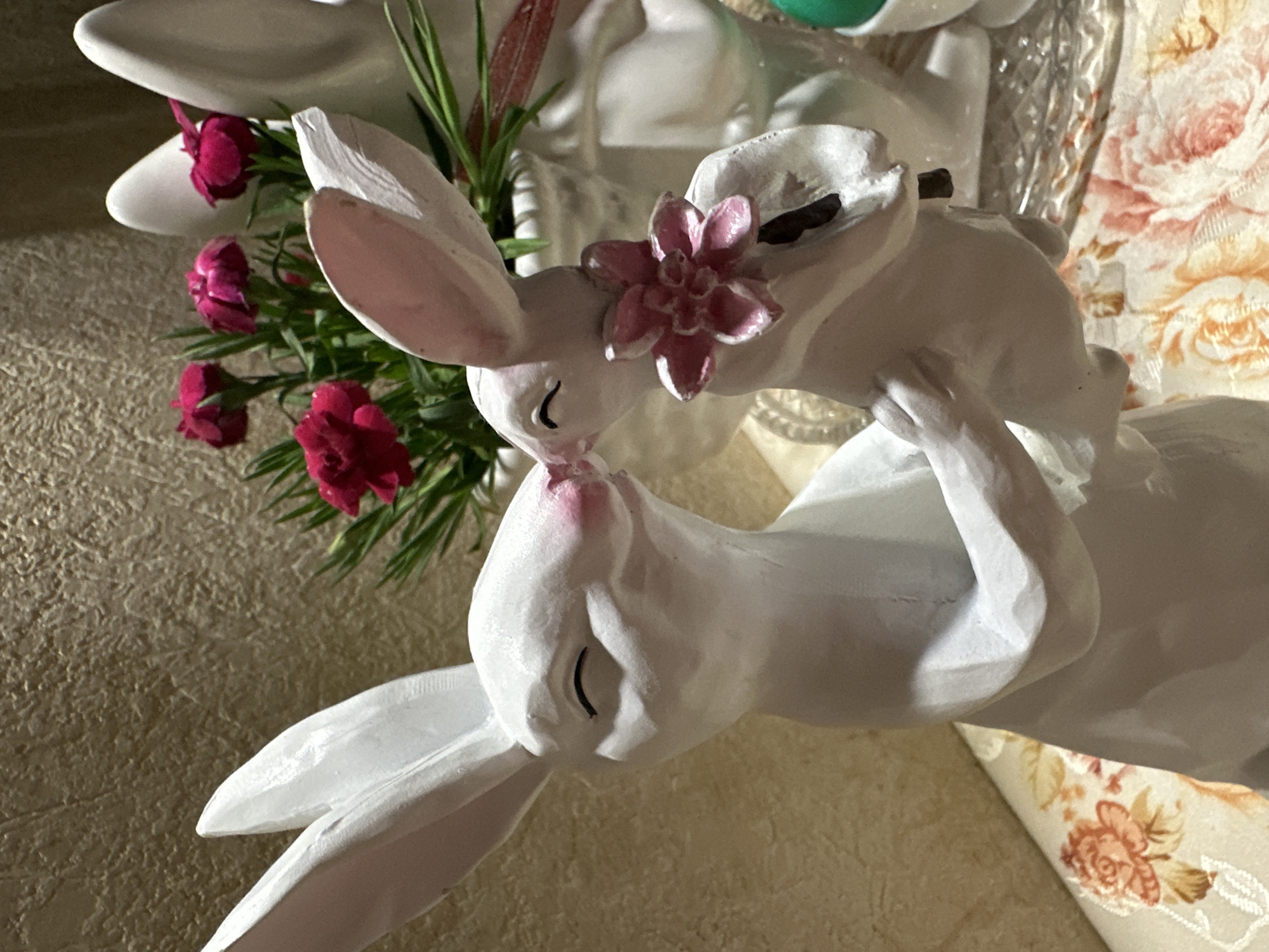 A close-up shot shows a porcelain figurine of two white rabbits kissing. The rabbits are detailed with painted eyes and small pink touches on their noses. A pink flower is positioned on the head of the top rabbit. Red carnations with green foliage are placed nearby. The rabbits sit on a surface that appears to be a floral fabric.