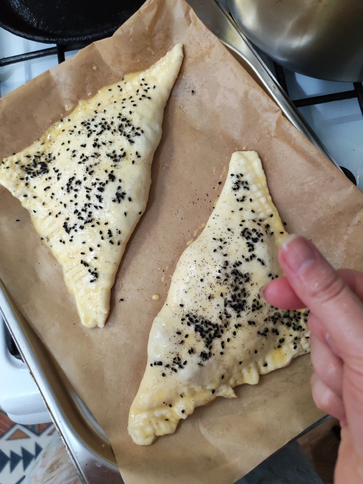 The image displays a close-up view of two unbaked, triangular pastries resting on a baking sheet covered with light brown parchment paper in a kitchen setting. The pastries appear to be brushed with an egg wash or oil, giving them a glossy sheen, and are liberally sprinkled with black sesame seeds, along with some finer, dark specks, possibly ground pepper or another spice. Their edges are distinctly crimped, suggesting they are filled and sealed.
A person's hand is visible on the right side of the frame, caught in the act of sprinkling additional seasoning onto the lower right pastry, indicating an ongoing food preparation activity.
In the background, elements of a domestic kitchen are discernible. A black pan, possibly cast iron, is partially visible in the upper left corner. Towards the upper right, a metallic pot or bowl and a pair of tongs can be seen. A white kitchen appliance or counter surface is also present at the edges of the frame. The scene portrays the final stages of preparing these items for baking in a kitchen, located in Izhevsk, Russia. The lighting suggests an indoor setting, but the specific time of day is not evident. No text is visible within the image.