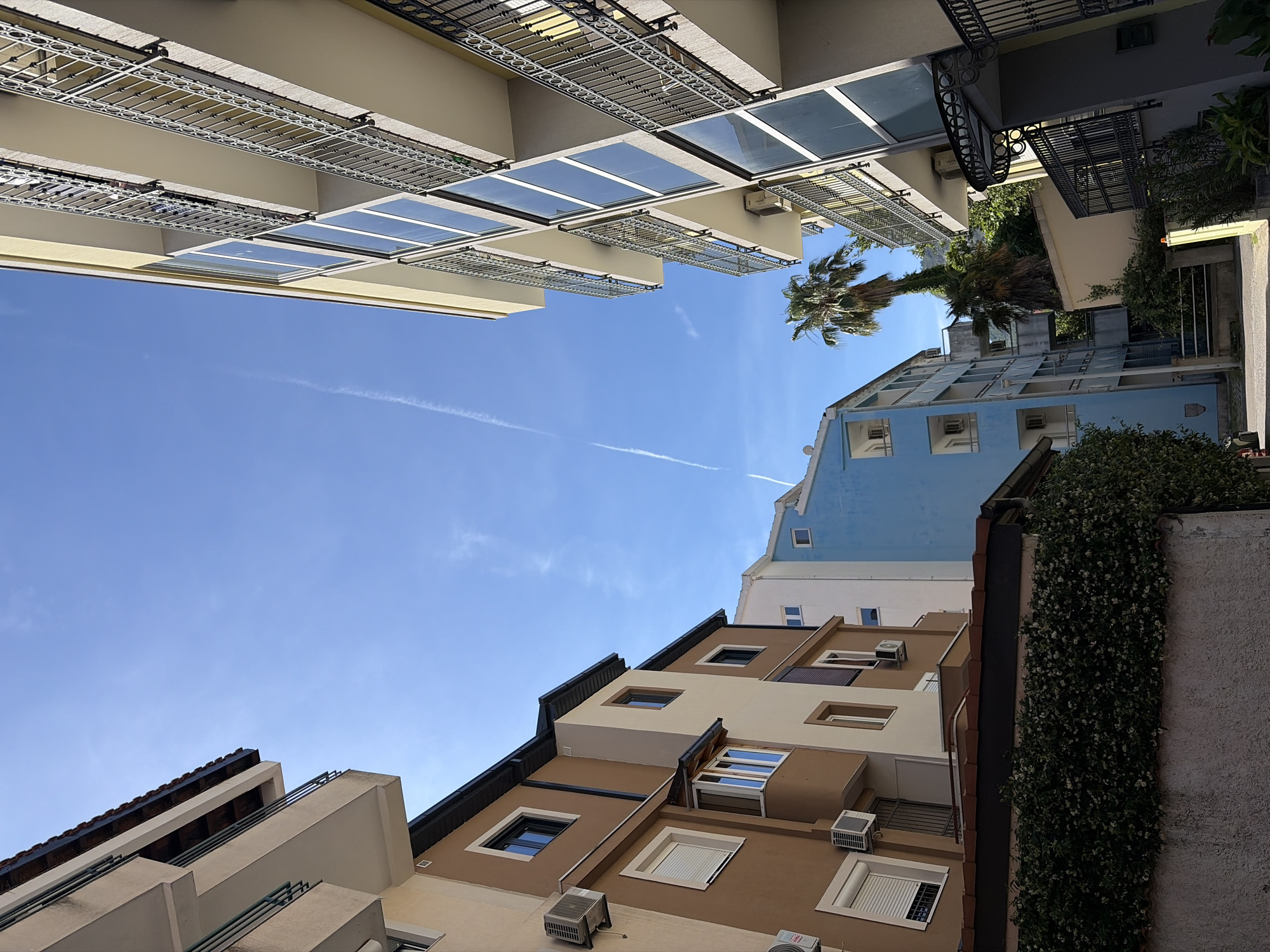 An upward-angled view within an urban area, likely in Budva, Montenegro, depicts buildings surrounding a narrow space with a bright blue sky overhead. The architecture is a mix of residential buildings with varying facades: balconies with ornate iron railings, light-colored structures with modern glass features, and buildings with earth-toned exteriors adorned with air conditioning units. A palm tree extends into the sky. A condensation trail suggests recent air travel. Greenery, including vines climbing on a wall, is visible.