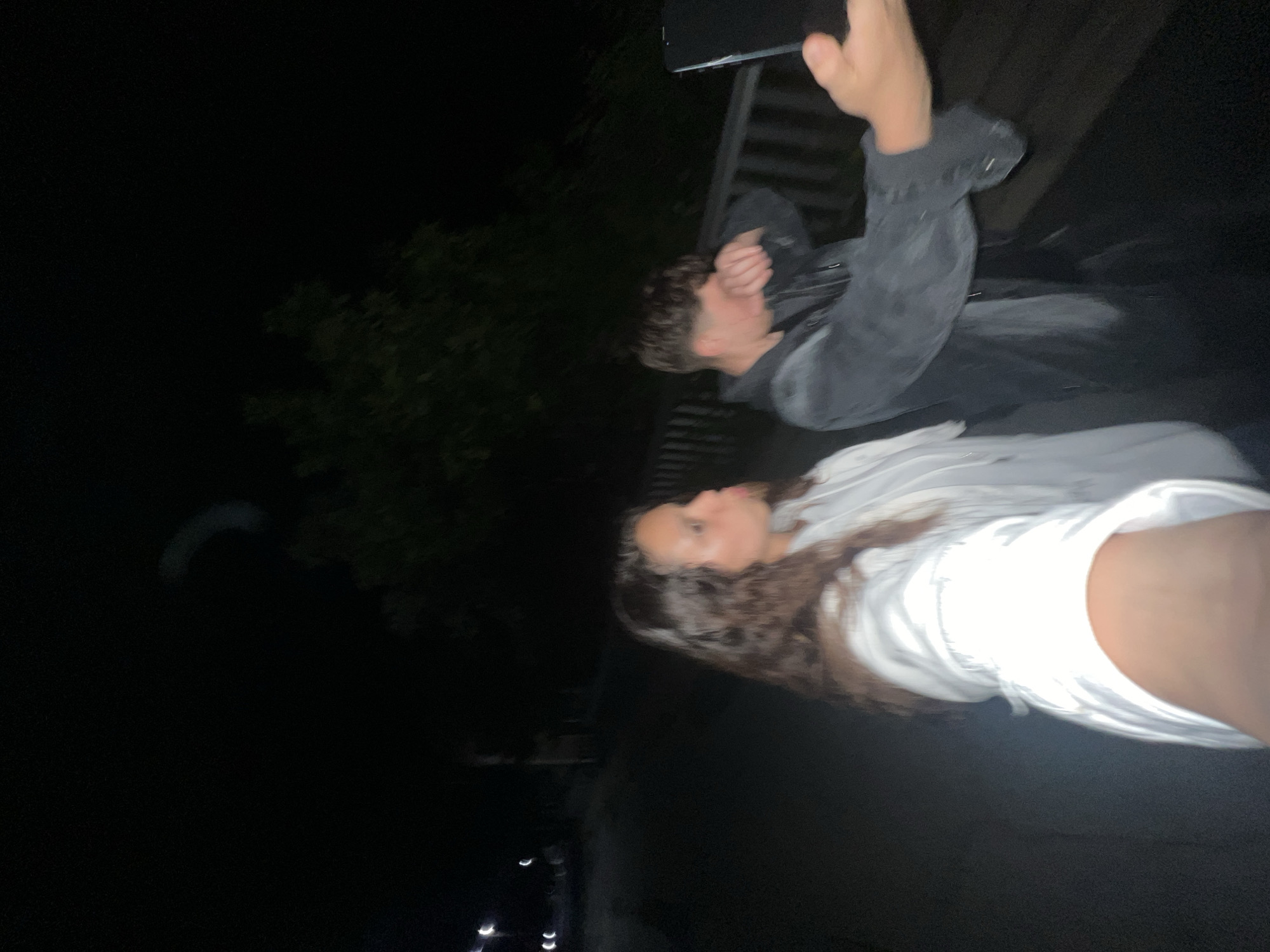 The image depicts two individuals outdoors at night in the city of Unknown, Unknown. The scene is underexposed and blurry, typical of low-light conditions.
In the foreground and midground, a female individual with long, dark, curly hair and a light-colored hooded sweatshirt extends her left arm towards the camera, suggesting she is holding the device. Her face is in profile, looking towards the right with pursed lips. To her right, a male individual with short, dark hair wears a dark jacket. His right hand is raised to his face, partially obscuring it, while his left hand holds a dark rectangular object, likely a smartphone, extended outwards. Both individuals appear to be posing for a photograph.
The background is dark. To the far left, multiple blurred light sources are visible, consistent with distant streetlights or vehicle lights along a road or path. To the right and behind the individuals, a dark wooden or metal railing structure is visible, suggesting a bridge, walkway, or elevated platform. In the upper right quadrant, dark shapes resembling trees are visible against the dark sky. A faint, light-colored, curved streak is present in the upper right sky, possibly a reflection or atmospheric phenomenon.