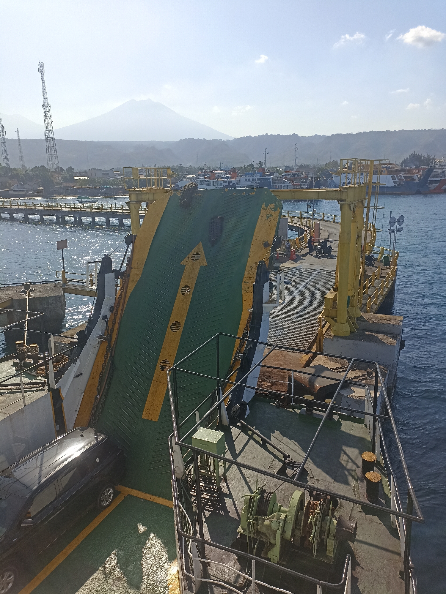 In Ketapang, Indonesia, a ferry ramp extends from a dock into the bright blue water. A black SUV is parked on the ramp, facing away from the camera. The ramp is painted green with yellow stripes and an arrow pointing forward, indicating the direction of travel. Behind the ramp, a yellow metal structure supports the dock, with stairs leading up and railings along the sides. Several motorcycles are parked on the dock, and people are visible walking around. In the background, a hazy mountain looms over a coastline dotted with buildings and trees. Several ferries are docked in the harbor, and two tall communication towers rise above the landscape. The sun casts strong shadows on the dock and ramp, indicating a clear, sunny day.