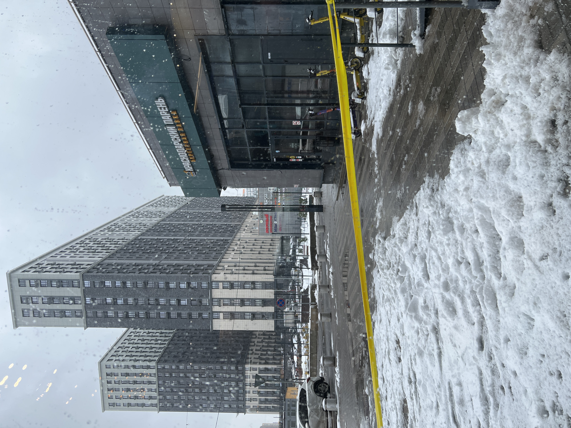 This image captures an urban street scene in Mytishchi, Russia, on an overcast winter day, likely viewed through a window with raindrops or snowflakes visible on its surface.
The primary subjects include a large, modern multi-story residential building on the left, featuring a distinct architectural design with varying shades of grey and numerous windows. A commercial building with a dark, glass-paneled facade stands on the right.
The street level features a paved surface, partially covered by significant accumulations of snow, particularly on the right side. A bright yellow line runs horizontally across the lower part of the frame, possibly indicating a lane or restricted area. Several shared mobility bicycles and scooters are parked near the commercial building, with some displaying the brand name "KICKSHARE." Further down the street, a white car is parked, and a row of street bollards is visible. A "No Stopping" traffic sign (red circle with a diagonal line on a blue background) is also present.
There is no discernible human activity taking place. The scene portrays a typical urban environment during cold weather.
Notable details include the heavy snow cover and the visible precipitation, indicating cold, winter conditions. The time of day appears to be daytime, but the sky is uniformly grey and overcast.
Visible text on the commercial building's facade reads:
- "КРАСНОДАРСКИЙ ПАРЕНЬ" (Krasnodarsky Paren, meaning "Krasnodar Guy")
- "ФИРМЕННАЯ ЛАВКА" (Firmennaya Lavka, meaning "Branded Shop" or "Company Store")