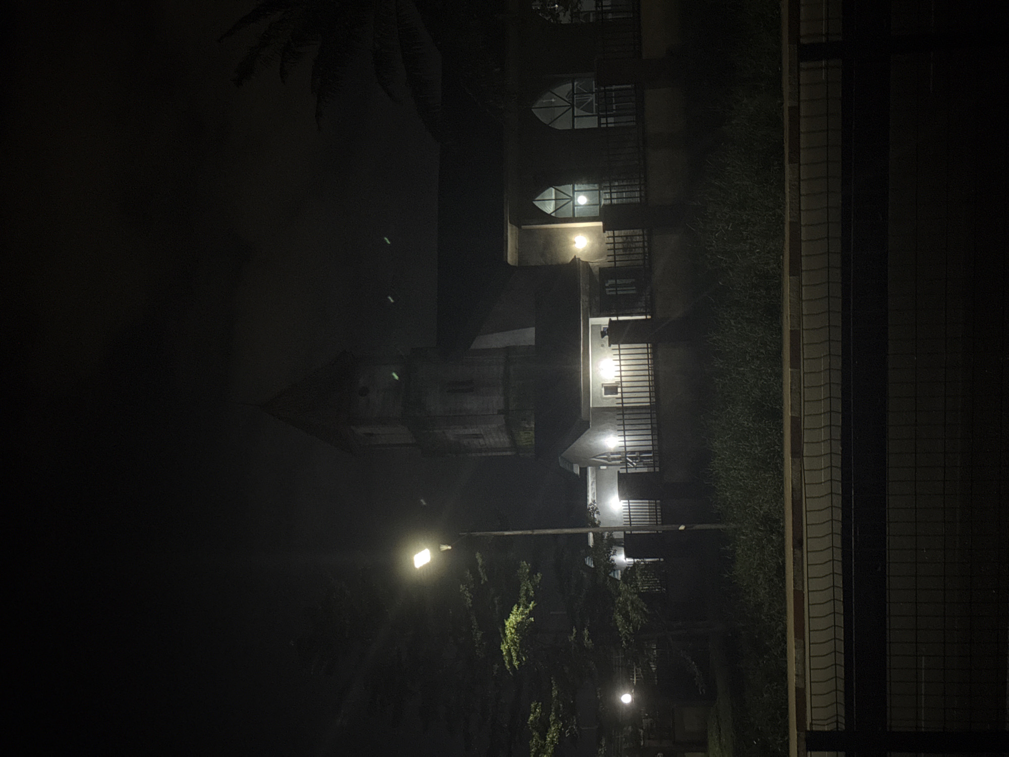 The image captures a dimly lit scene at night, presumably in Uyo, Nigeria, featuring a church building. A tall street lamp illuminates the foreground, casting light on a nearby tree with green foliage and a dark, wrought-iron fence. The church itself is a multi-story structure with a prominent tower, and its windows are illuminated from within, suggesting occupancy. The building's architecture appears to be traditional, with arched windows visible on the main facade. Adjacent to the church is a lower, modern-looking section, also with illuminated windows and a fence. Beyond the fence, dense, dark foliage suggests a natural, untamed area. In the foreground, a paved road with a curb and a black metal fence are visible, indicating a public thoroughfare. The overall atmosphere is serene and slightly mysterious, with the darkness of the night enveloping the scene.