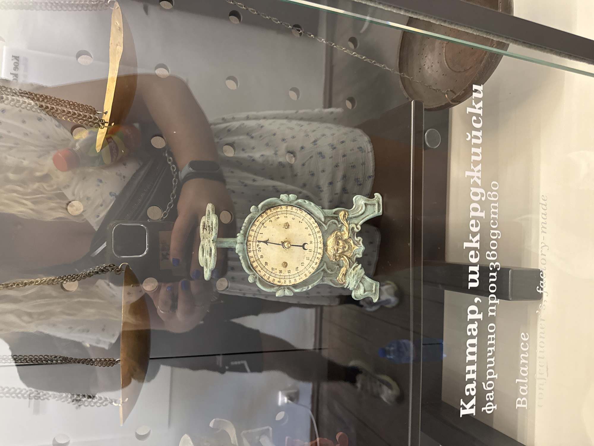 The image shows a close-up of a glass display case containing an antique balance scale. The scale is ornate, made of a greenish-bronze metal, and has a circular dial with Roman numerals. The dial is marked with various weights and measurements, including "Kilo 0". A person, partially reflected in the glass, is holding a smartphone and appears to be taking a picture of the display. The person is wearing a white dress with a blue floral pattern and a dark fanny pack. They have blonde hair and are wearing rings on their fingers. The background shows a reflection of another person standing in shorts and sneakers. Text in Bulgarian and English is visible on the glass, identifying the item as a "Balance, confectioner's, factory-made". This display is located in the Museum of Crafts and Applied Arts in Gabrovo, Bulgaria.