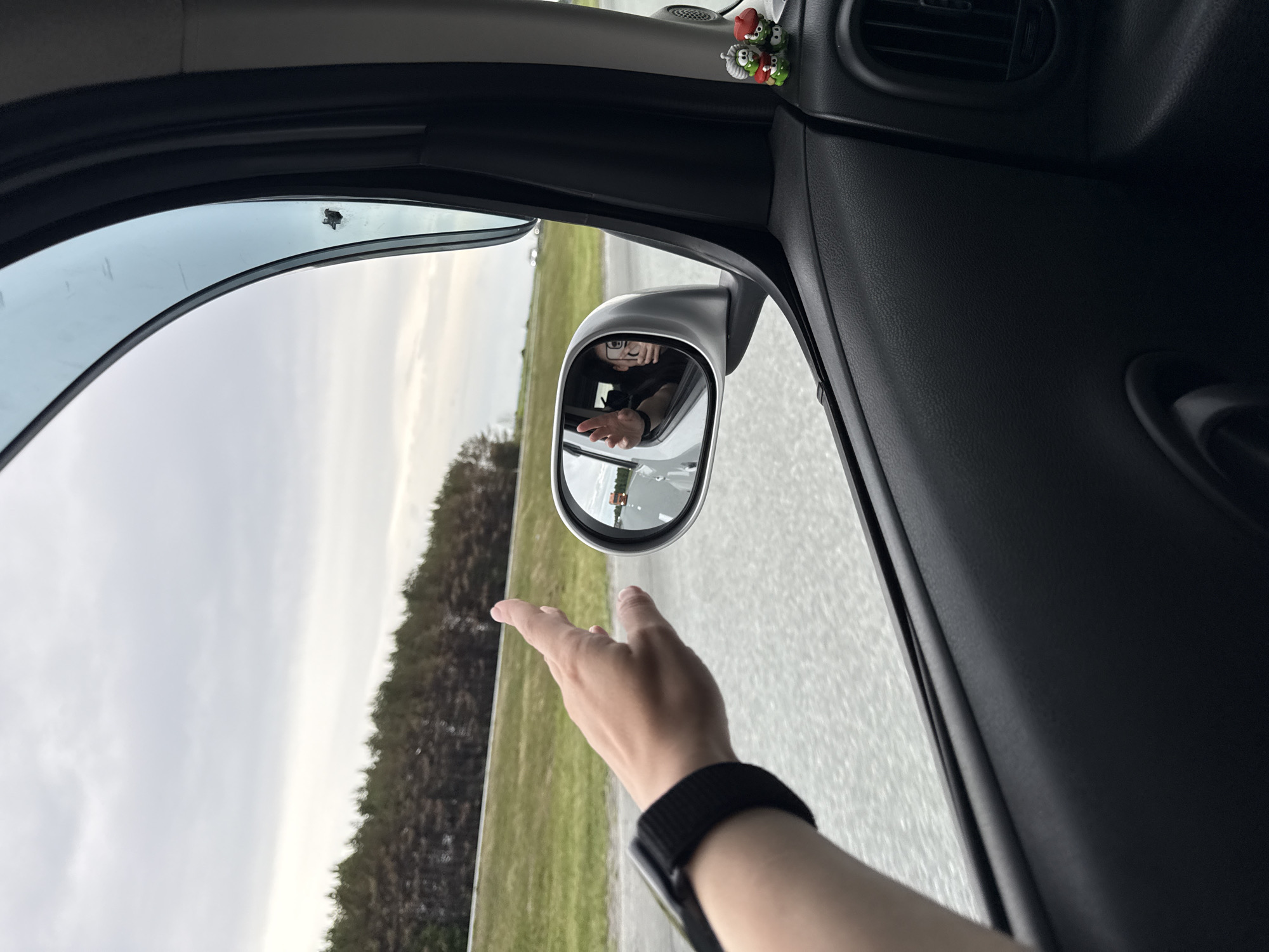The image, taken from inside a car, shows an arm extending out of the window. The wrist has a black watch band. The hand appears to be waving. The side mirror reflects a person wearing glasses, also waving. Outside, the landscape consists of a road flanked by a field or grassy area and a treeline. The sky is overcast. In the car's interior, a small decorative object with multiple figures is attached to the car's dashboard. The city is not identifiable in this image.