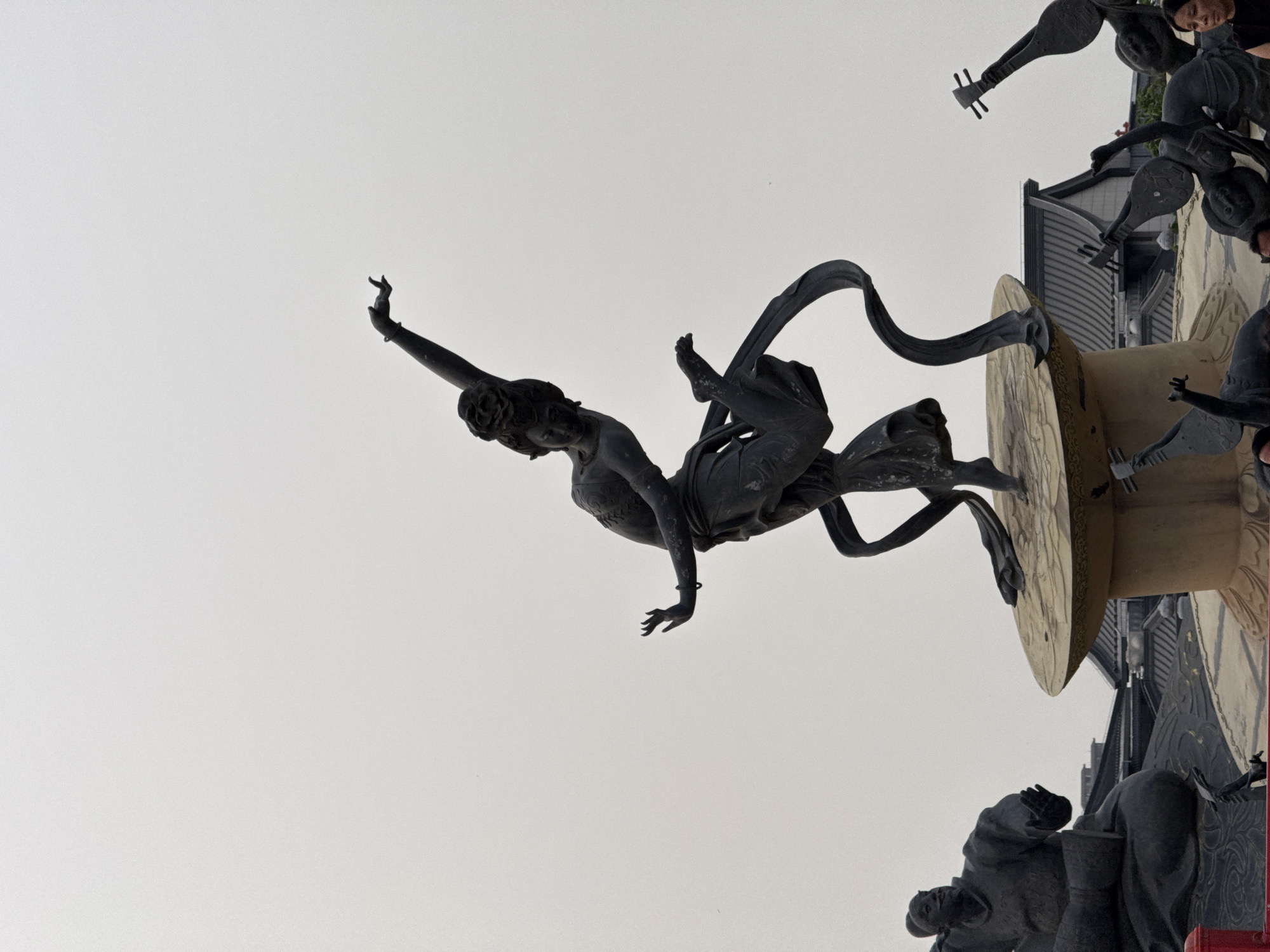 This image shows a statue in Lintong District, China. A bronze statue of a female figure, in mid-air, is supported by a circular base. The figure is posed in a dance-like posture with one arm raised, wrapped in flowing fabric. Around the base of the central figure are other smaller statues playing stringed instruments, with Chinese architecture visible in the background. The sky overhead is overcast.