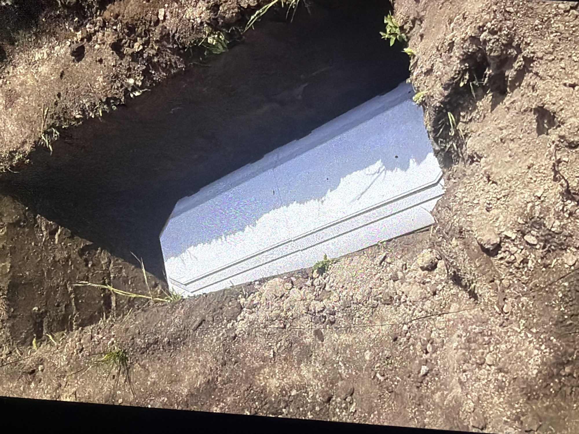 A light-colored, rectangular object, possibly a casket, is partially visible within a freshly dug grave. The grave is a rectangular pit in the earth, with soil piled up around its edges. Some sparse green vegetation is growing in the soil. The scene appears to be outdoors in Bukuru, Nigeria, as indicated by the surrounding environment and soil type.
