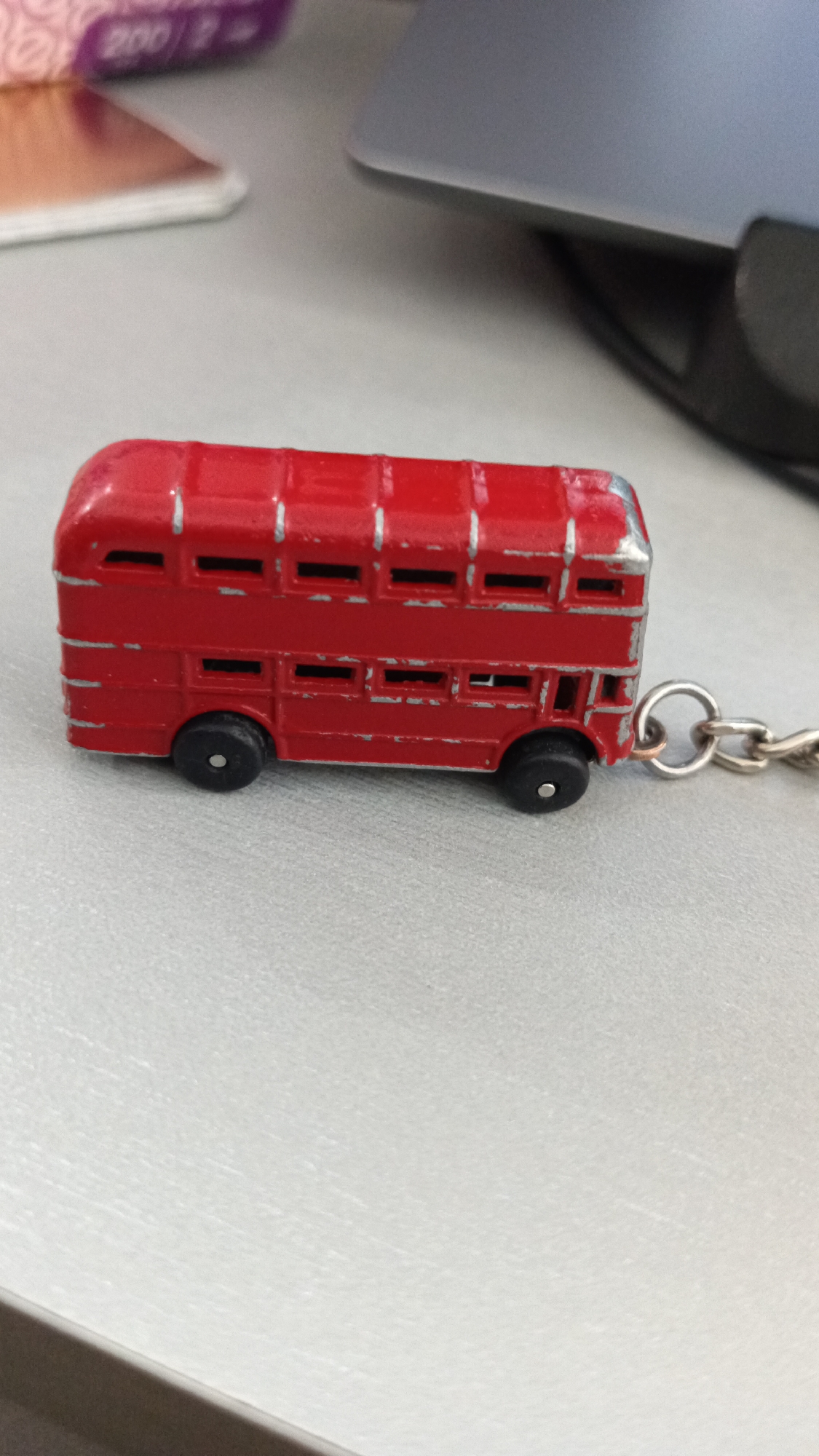 This media file features a close-up, eye-level shot of a red double-decker bus toy, likely a miniature replica of a London bus. The toy is a keychain, with a silver chain and a ring attached to its rear. The bus itself is painted a bright red with silver trim around the windows and along the edges of its body. It has two visible levels of windows, all of which appear to be dark, indicating they are either tinted or blacked out. Two black wheels are visible on the lower level, suggesting it's a toy vehicle. The toy is resting on a light-colored, textured surface, possibly a desk or table. In the blurred background, a silver laptop is partially visible on the right side, and a portion of a tissue box or similar item with purple and pink markings is visible on the left. The lighting suggests an indoor environment, possibly during daylight hours. There are no people or overt activities depicted; the focus is solely on the toy bus. The overall impression is that of a personal item, perhaps a desk accessory or a souvenir, placed on a work surface. The environment appears to be a casual setting.