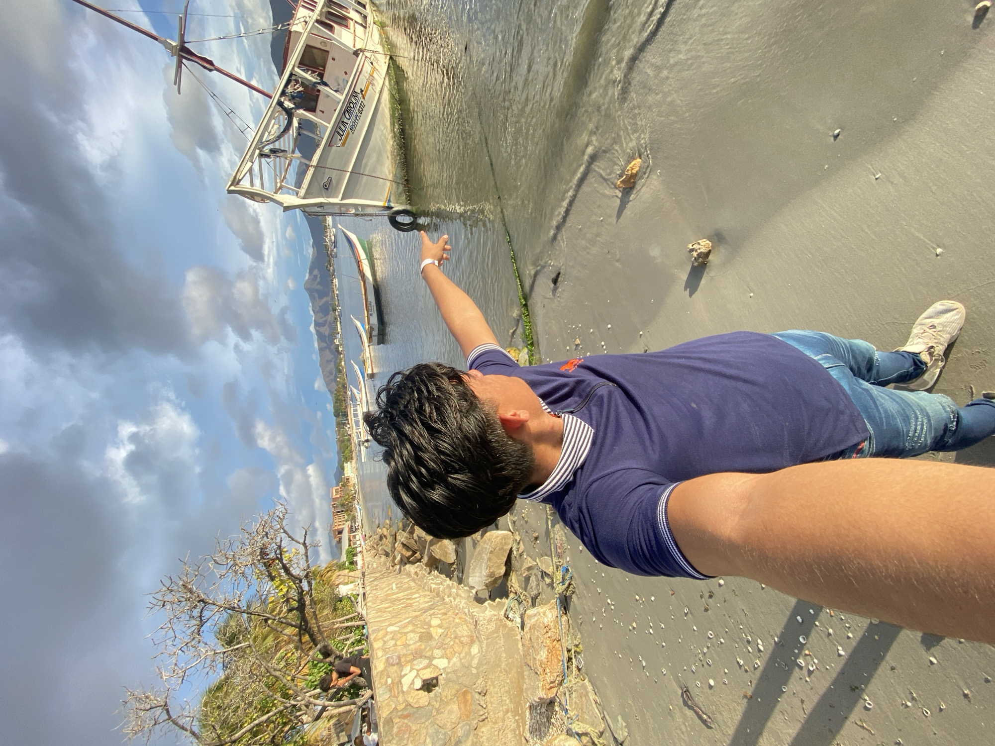 A man is taking a selfie on a beach in Juan Griego, Venezuela. The man is wearing a blue shirt and jeans, and he is pointing towards a beached boat. The tide is coming in and lapping at the sand near the boat. The sky is cloudy. In the background there are mountains and buildings. There is a stone wall and a bare tree next to the man, and two people lying under the tree.