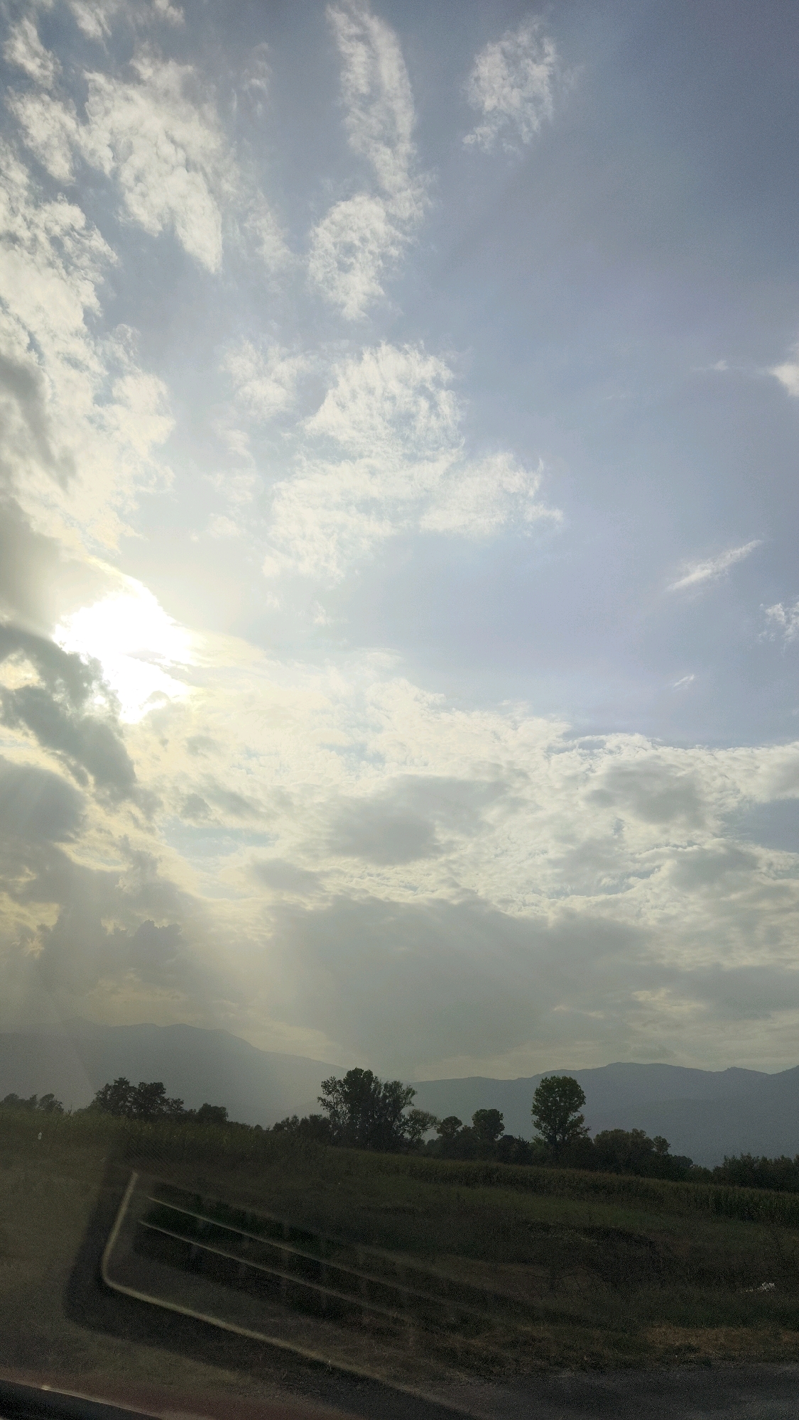 The image shows a cloudy sky over a rural landscape. In the foreground, there is a grassy field and some trees. In the distance, there are mountains. The sun is visible through the clouds, casting a soft light on the scene. The location is identified as Shemshevo, North Macedonia. There are no people or animals visible in the image.