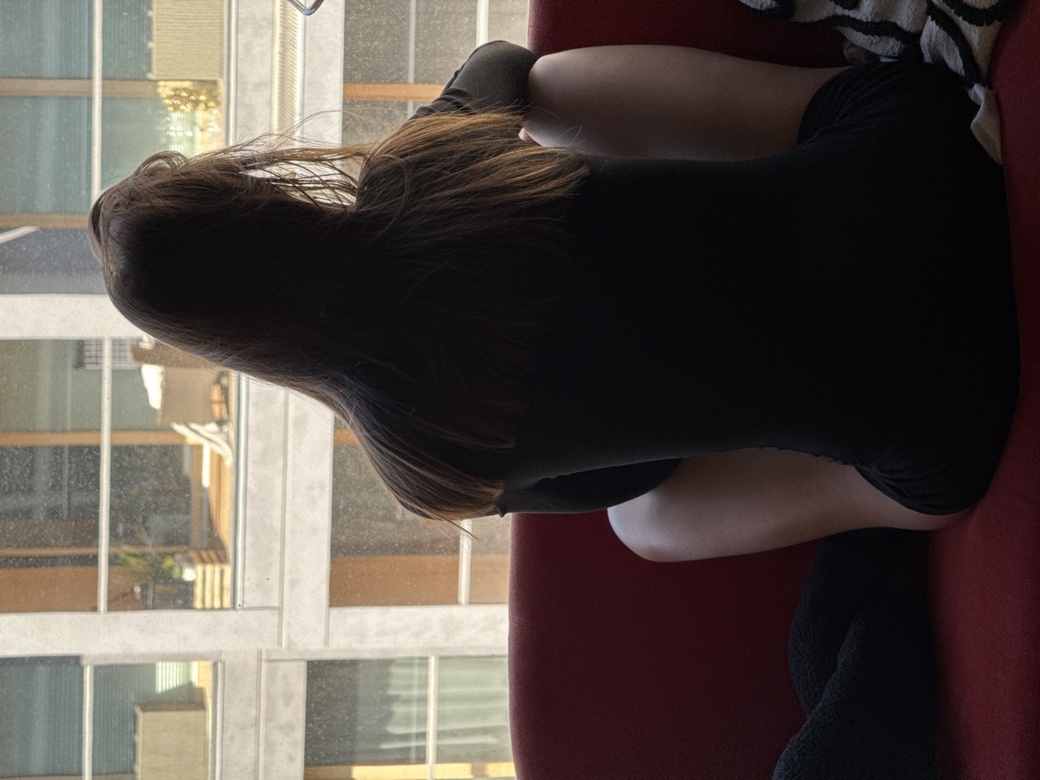 The image, when viewed upright, depicts a person from behind, seated or kneeling on a red sofa or cushion, facing a large window. The individual has long, dark brown hair that falls over their back and shoulders, and they are wearing a plain black, form-fitting outfit.
The person appears to be looking out the window, which offers a clear view of an urban environment. Visible through the window is the facade of a modern building, featuring a grid of rectangular windows. Some of these windows reflect bright daylight, indicating it is daytime, while others reveal interior details like blinds or ambiguous shapes. The indoor area where the person is situated is somewhat shadowed, creating a silhouette effect that emphasizes the person's form and hair against the brighter outdoor scene.
The activity suggests a moment of quiet observation or contemplation. The setting is an indoor space, likely an apartment or room, offering a perspective on the city of Warsaw, Poland, as indicated by the location context. There is no visible text in the image.