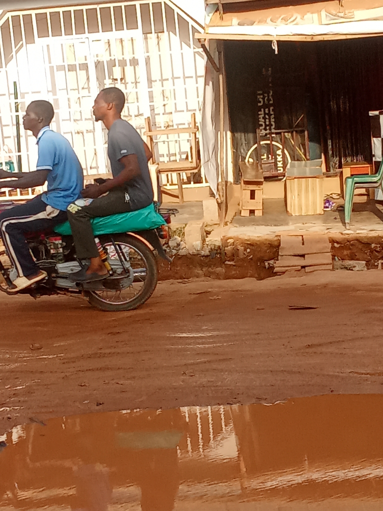 The image depicts two young men on a motorcycle on a dirt road in Kagini, Nigeria. The man in front is wearing a blue t-shirt and dark pants, while the man behind him is wearing a gray t-shirt and dark pants. They are both looking forward. The motorcycle is red with a green seat cover. The background shows a building with a white metal facade and a dark curtained shop entrance. There are some wooden shelves and chairs outside the shop. The road is muddy and there are puddles of water on the ground. The lighting suggests it is daytime.