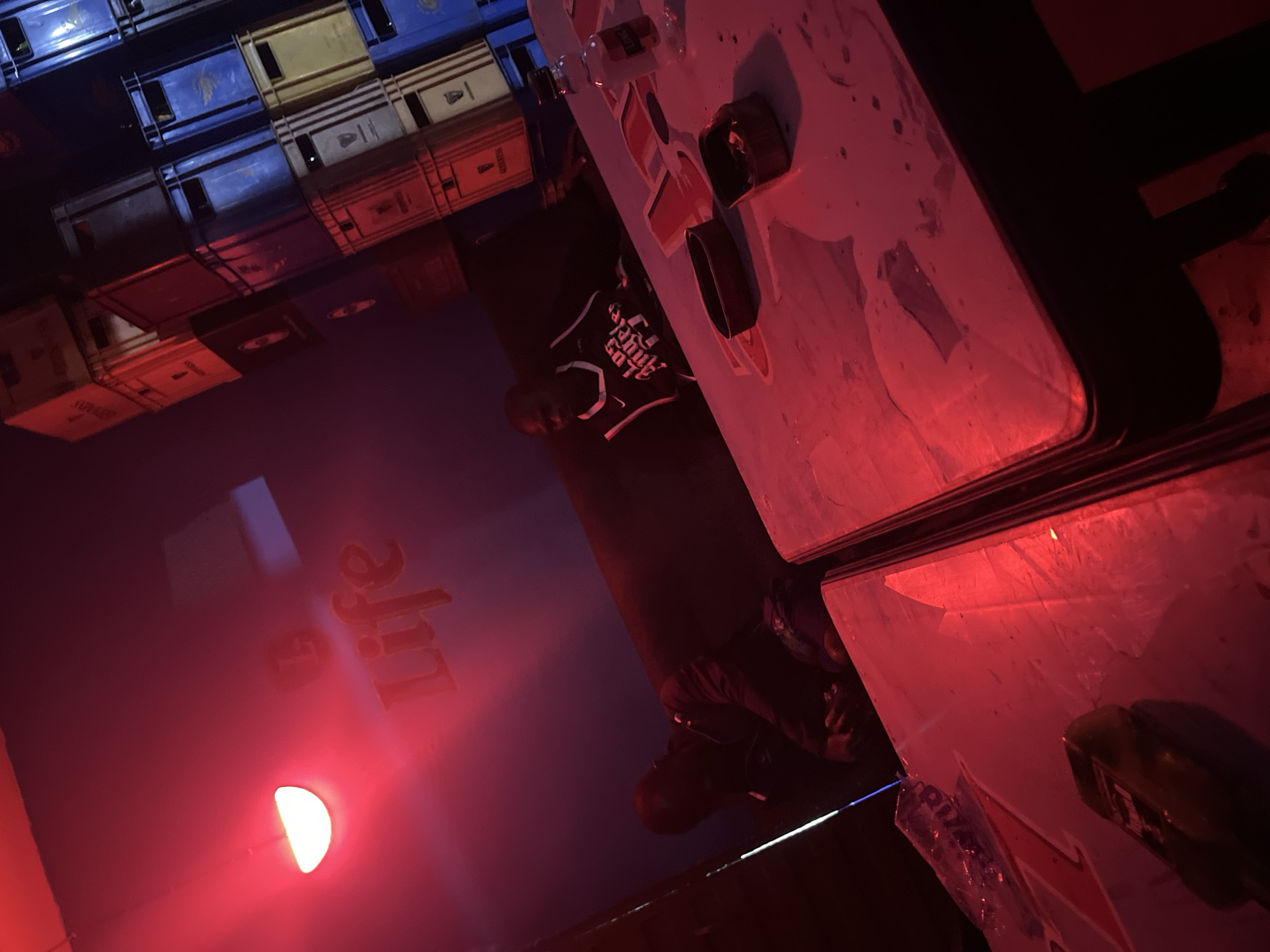 In a dimly lit indoor setting with red lighting, the scene appears to be a bar or club in Jos, Nigeria. The background features stacks of colorful crates, possibly for beverages. In the foreground, a table is visible, with two ashtrays and a bottle of liquor. Two individuals are present: one man is seated at the table, while another is partially visible in the background, hunched over and seemingly engaged in conversation or activity. The overall ambiance suggests a relaxed or possibly clandestine atmosphere.