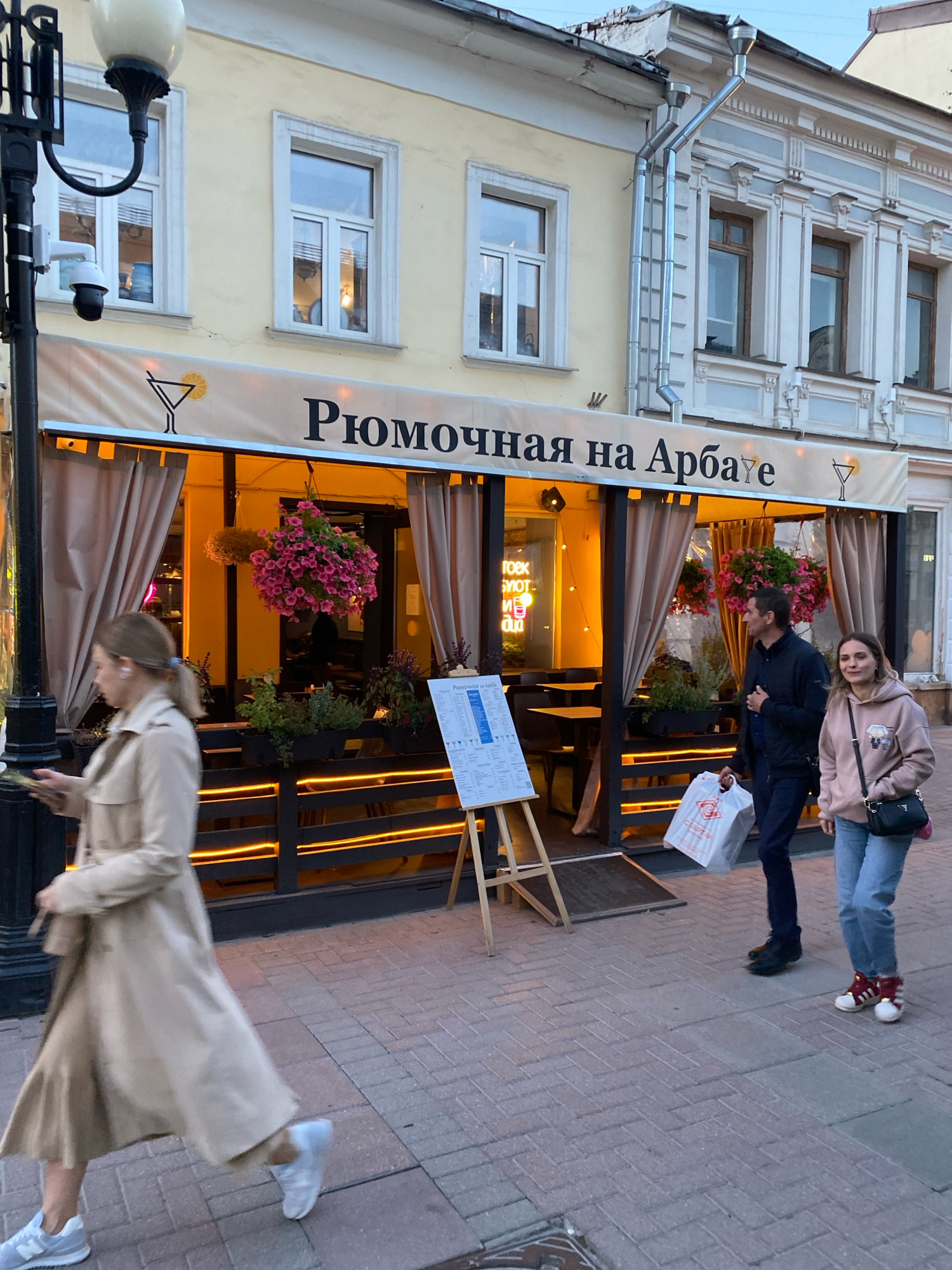 An exterior, eye-level shot captures a street scene in Moscow, Russia, featuring a restaurant with Cyrillic signage that reads "Рюмочная на Арбате". The establishment has a light yellow facade with large windows, partially obscured by beige drapes. Hanging floral arrangements, one a vibrant pink, add a touch of color. The restaurant is brightly lit from within, with warm yellow lights illuminating the interior and an orange glow along the exterior railing. Two individuals, a man and a woman, are walking past the restaurant on a brick-paved sidewalk. The man, dressed in a dark jacket and pants, carries a white shopping bag. The woman, wearing a pink hoodie and jeans, has a black purse slung over her shoulder. To the left of the restaurant, a woman in a beige trench coat is walking away from the camera, holding a phone. A black lamppost with a security camera is visible on the far left. A menu board stands on an easel in front of the restaurant. The overall lighting suggests it is late afternoon or early evening.