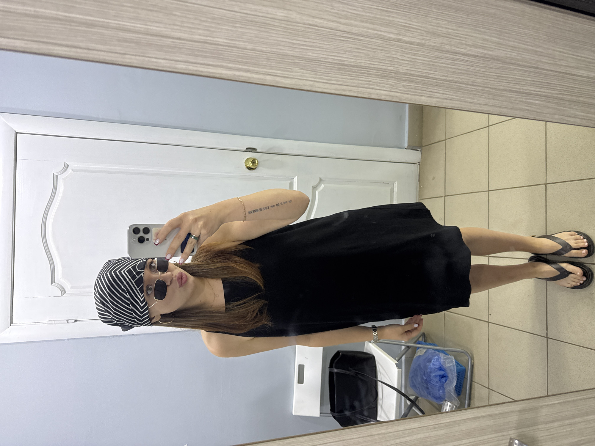 The image shows a young woman in a black dress and a black and white striped bandana, taking a mirror selfie. She is wearing sunglasses and flip-flops. Her left arm has a tattoo. She is in what appears to be a room with tiled floors and a white door. A small metal cart with a blue plastic bag is visible in the background. The lighting suggests it is daytime.