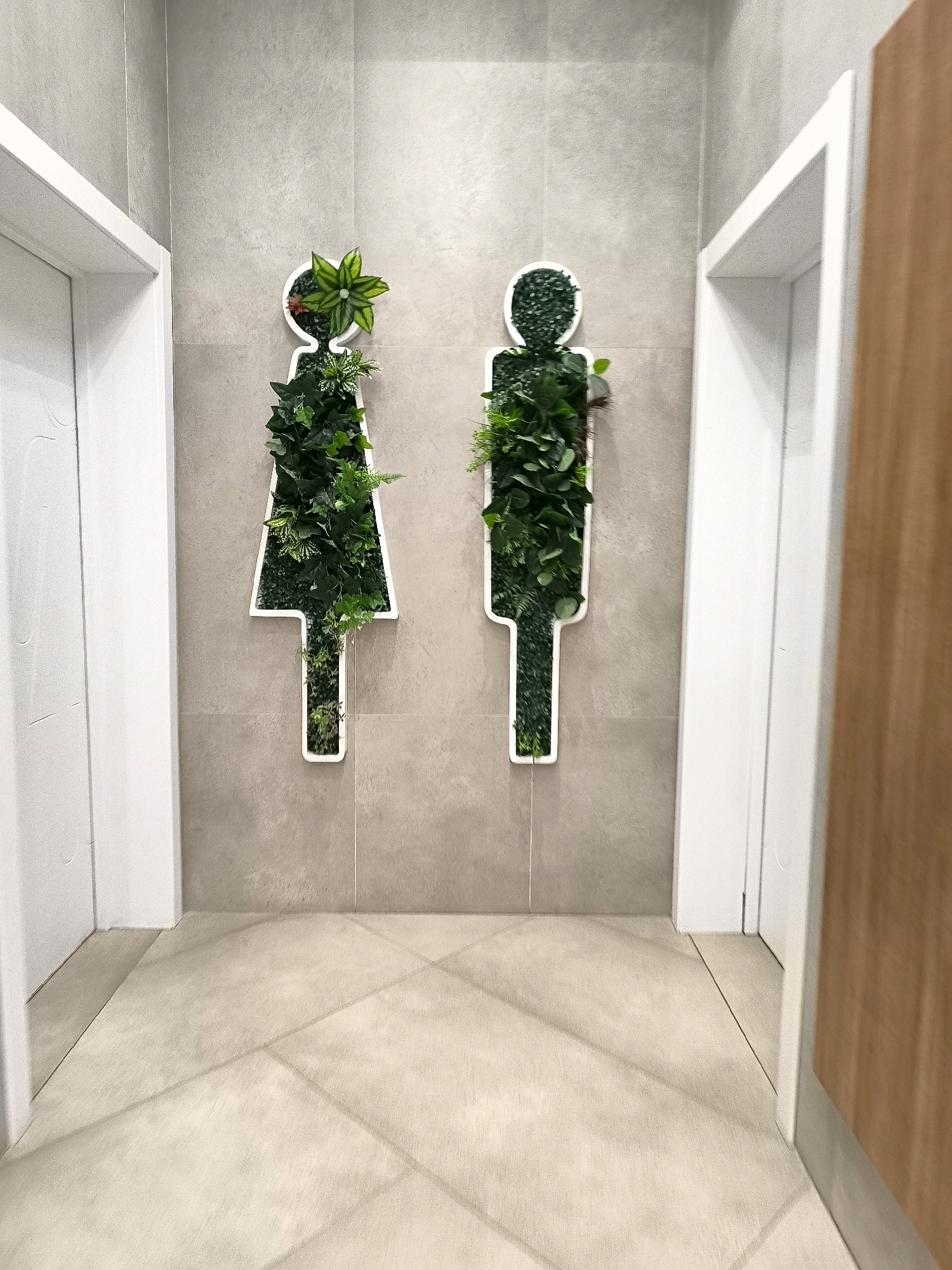 In the city administration of Qarağandı, Kazakhstan, two doors stand on either side of a grey tiled wall. Mounted on the wall are two stylized human figures, one on the left representing a female and the other on the right representing a male. These figures are outlined in white and filled with lush green artificial plants, creating a unique and decorative display. The floor is tiled with large, light-colored square tiles, laid out in a grid pattern, with subtle shadows cast from an unseen light source. The overall scene suggests a modern and possibly eco-conscious design within a public building.
