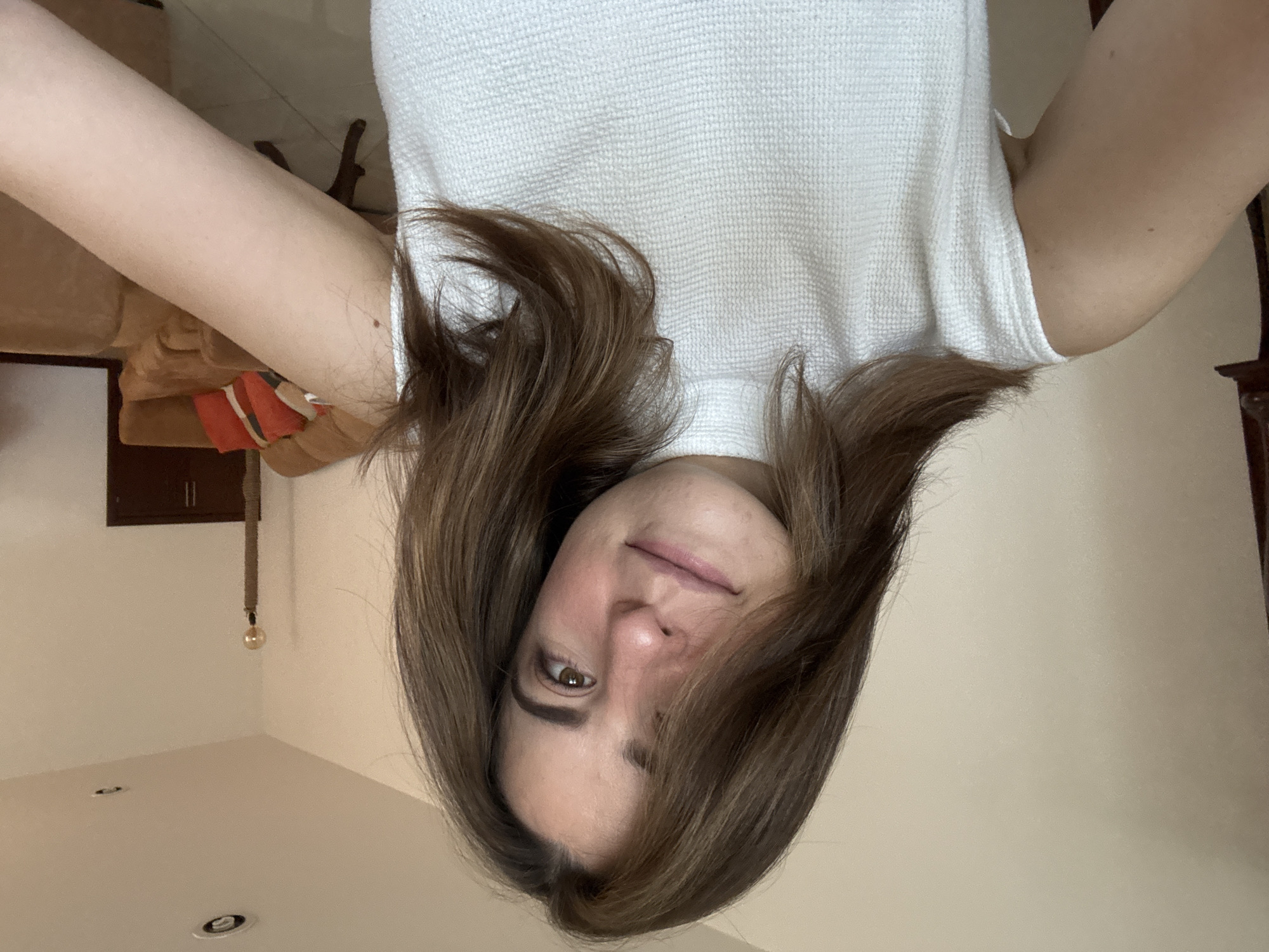 This media file is an upside-down selfie of a person in an indoor setting.
**Subjects:** The primary subject is a female with light skin, brown hair, and brown eyes. Her hair is shoulder-length, parted, and frames her face. She is looking directly at the camera with a subtle, closed-mouth smile.
**Objects:** She is wearing a white, textured top, possibly a waffle-knit fabric. In the background, visible above her head (which would be the floor if the image were upright), are light-colored walls or ceiling with two recessed lights or vents. A single light bulb hangs from a braided rope cord from what appears to be a dark brown cabinet or shelf attached to the wall. To her right in the image (the left side of the room in an upright view) is a partial view of a light brown or tan upholstered sofa, with a glimpse of an orange and white patterned item, possibly a throw pillow or blanket. On the opposite side, a dark wooden structure, potentially part of furniture or architectural detail, is visible.
**Scene/Setting:** The scene is an interior room, likely a living space or bedroom, characterized by light-colored walls and comfortable furniture. The known location context is Puebla, Mexico.
**Activity or event taking place:** The person is taking a selfie, looking directly into the camera.
**Notable details:** The image is distinctly presented upside down. The lighting appears bright and natural for an indoor setting during the daytime. The person exhibits a calm and pleasant expression. There is no visible text in the image.