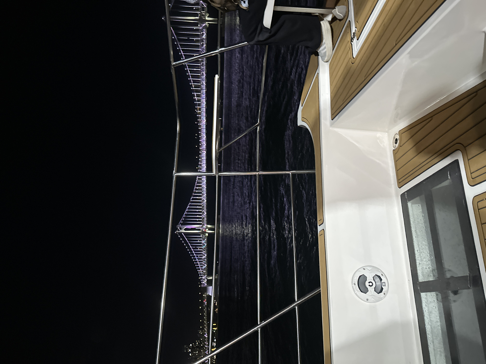 The image shows a view from a boat at night, looking towards a brightly lit suspension bridge and the Busan cityscape. The boat has white fiberglass hull with wooden decking. A person's legs are visible on the right side of the frame, wearing black pants and white sneakers, seated on the boat. The bridge, illuminated with purple and white lights, spans across a dark body of water, likely the sea or a bay, with the lights of the city stretching along the coastline in the background. The overall scene is one of a nighttime excursion on the water, with a prominent view of the Gwangan Diamond Bridge in Busan, South Korea.