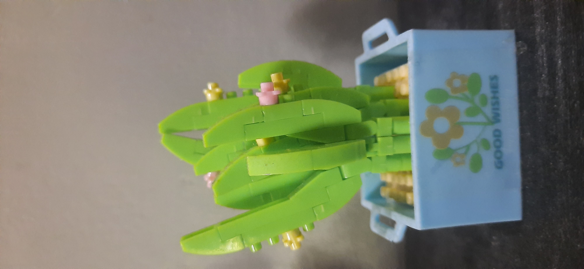 The image shows a close-up, eye-level view of a small, light blue plastic pot containing a miniature, artificial cactus plant. The pot has a decorative floral design on its front, featuring two yellow flowers with green leaves, and the words "GOOD WISHES" printed in dark blue below the flowers. The cactus is constructed from green plastic building blocks and has several small pink and yellow flowers attached. The pot is sitting on a dark, textured surface, likely a table or shelf. The background is a plain, light gray wall. The lighting appears to be from above and slightly to the front. There is no indication of the city of Cisauk, Indonesia, or any specific location within it.