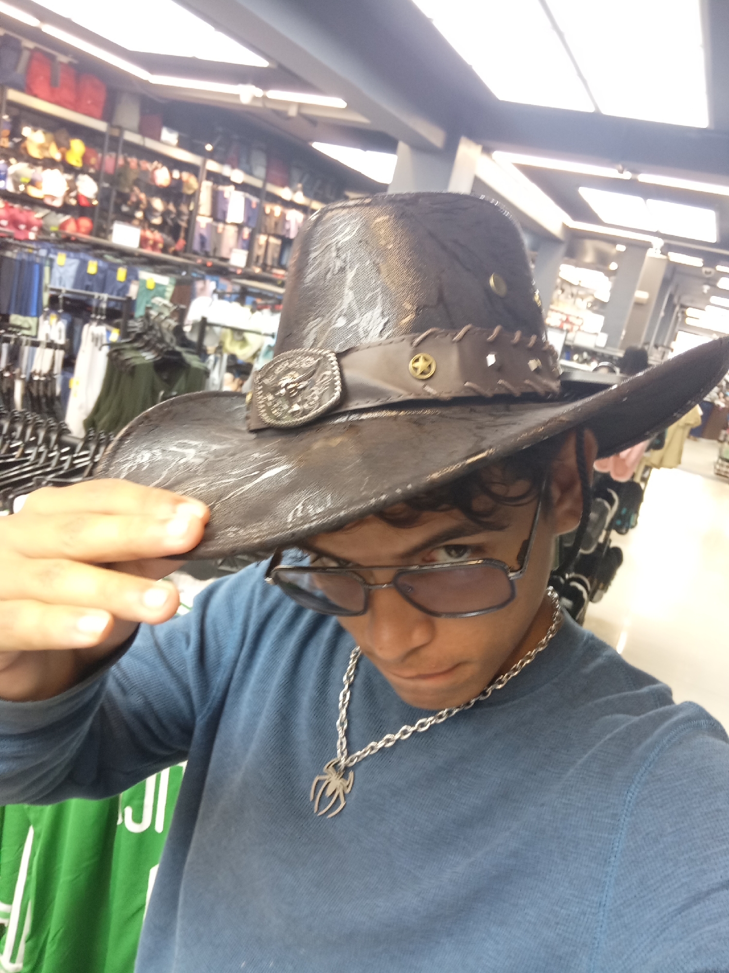 An individual, appearing to be a young male, is captured in a close-up, chest-up selfie in an indoor retail environment. He wears a dark, textured, wide-brimmed cowboy-style hat. The hat features a lighter brown decorative band with punched-out shapes, metallic studs, and a prominent silver-colored buckle on the front depicting an intricate design, possibly a bull or skull motif. He is wearing rectangular metal-framed glasses with dark-tinted lenses. His attire consists of a long-sleeved, light blue thermal-knit shirt and a silver-colored chain necklace with a distinct spider-shaped pendant. His left hand is partially visible, with fingers resting on the brim of the hat.
The background shows a retail store setting, with multiple clothing racks laden with garments on hangers. On the left, racks display numerous dark-colored shirts and pants, along with a partially visible green garment featuring white Arabic script (reading "الج" from right to left). In the upper left background, shelves hold various out-of-focus items, including what appear to be bags or accessories in red, yellow, and other colors. The store ceiling is dark grey or blue and is equipped with multiple bright, rectangular fluorescent light fixtures providing overhead illumination.