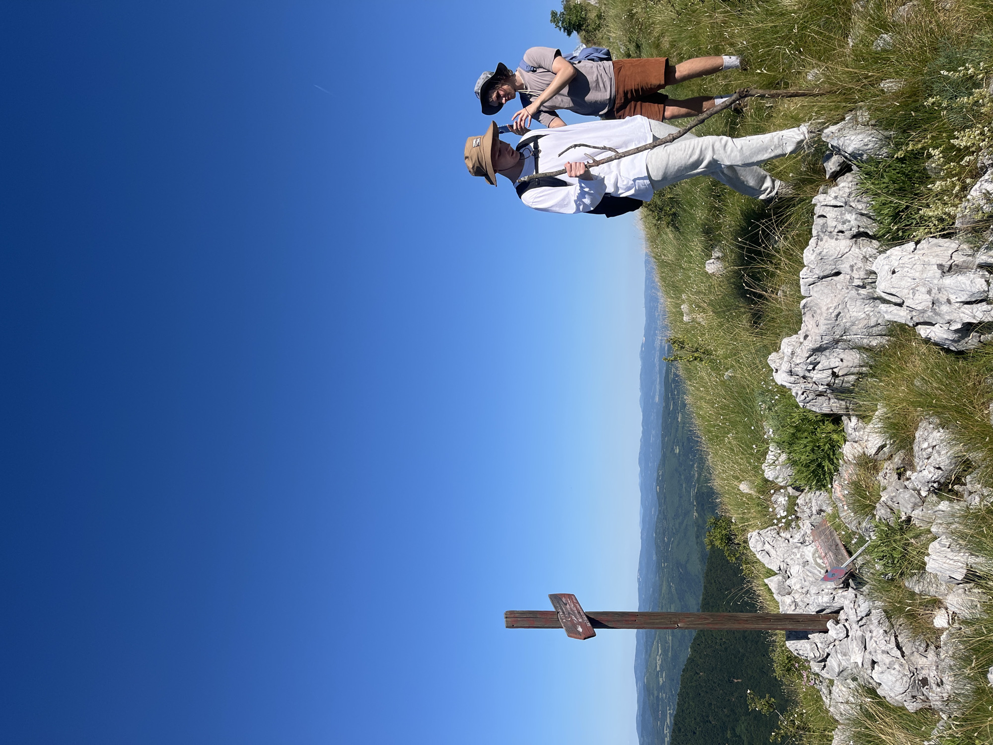 Two young males are hiking on a grassy, rocky hillside under a clear blue sky. The person in the foreground is wearing a white long-sleeved shirt, light grey pants, a light brown bucket hat, and a backpack. They are holding a long, thin branch as a walking stick. The person behind them is wearing a grey t-shirt, brown shorts, a patterned bucket hat, and a blue backpack. They are holding up a smartphone, seemingly taking a photo. A wooden signpost with two directional signs is to the left of the foreground hiker. The background shows rolling hills and a distant mountain range. This scene is captured in the vicinity of Petrovac-na-Mlavi Municipality, Serbia, suggesting a hiking trail or viewpoint in a mountainous or hilly terrain.
