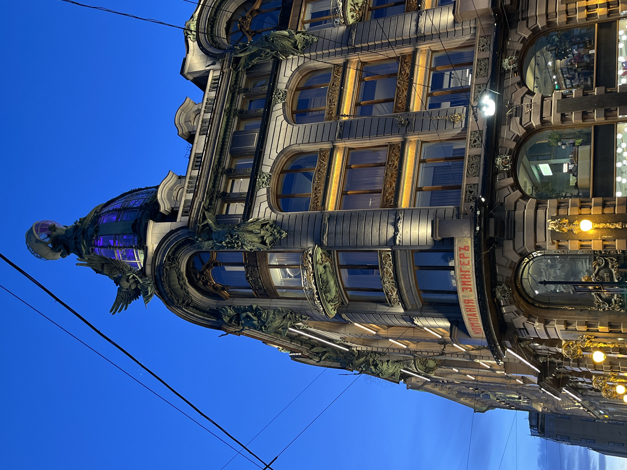 The image shows the Singer House, an iconic building in Saint Petersburg, Russia. The building is illuminated with warm, golden light highlighting its ornate details, including decorative carvings, arched windows, and sculptures. A glass dome, topped with a statue, is visible on the corner of the building. The sky is a deep blue. A sign with Cyrillic text is visible on the building's facade. Overhead wires are present in the sky.
