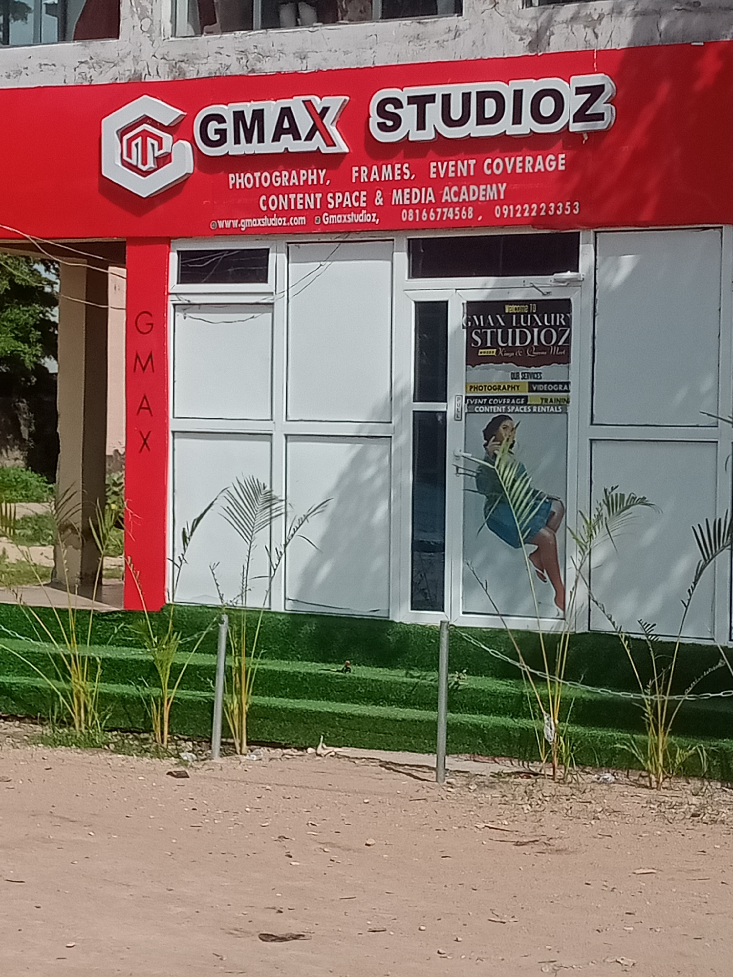 The image shows the exterior of a business named "GMAX STUDIOZ" in Jalingo, Nigeria. The business is located in a building with a prominent red facade that features the business name in large, white, 3D lettering. Below the business name, smaller white text lists services such as "PHOTOGRAPHY, FRAMES, EVENT COVERAGE CONTENT SPACE & MEDIA ACADEMY". Contact information, including a website address and phone numbers, is also visible.
The entrance to the studio is a glass door with a sign that reads "WELCOME TO GMAX LUXURY STUDIOZ". The sign also lists "OUR SERVICES" including "PHOTOGRAPHY, VIDEOGRAPHY, EVENT COVERAGE, TRAINING, CONTENT SPACES RENTALS". A stylized image of a woman is visible on the glass door.
The building has large white-framed windows. In front of the studio, there is a strip of artificial green turf with several potted plants and palm fronds. The ground in front of the turf is a dirt path with small stones and debris. The surrounding environment appears to be an urban or semi-urban setting, with other buildings and vegetation visible in the background. The lighting suggests it is daytime.