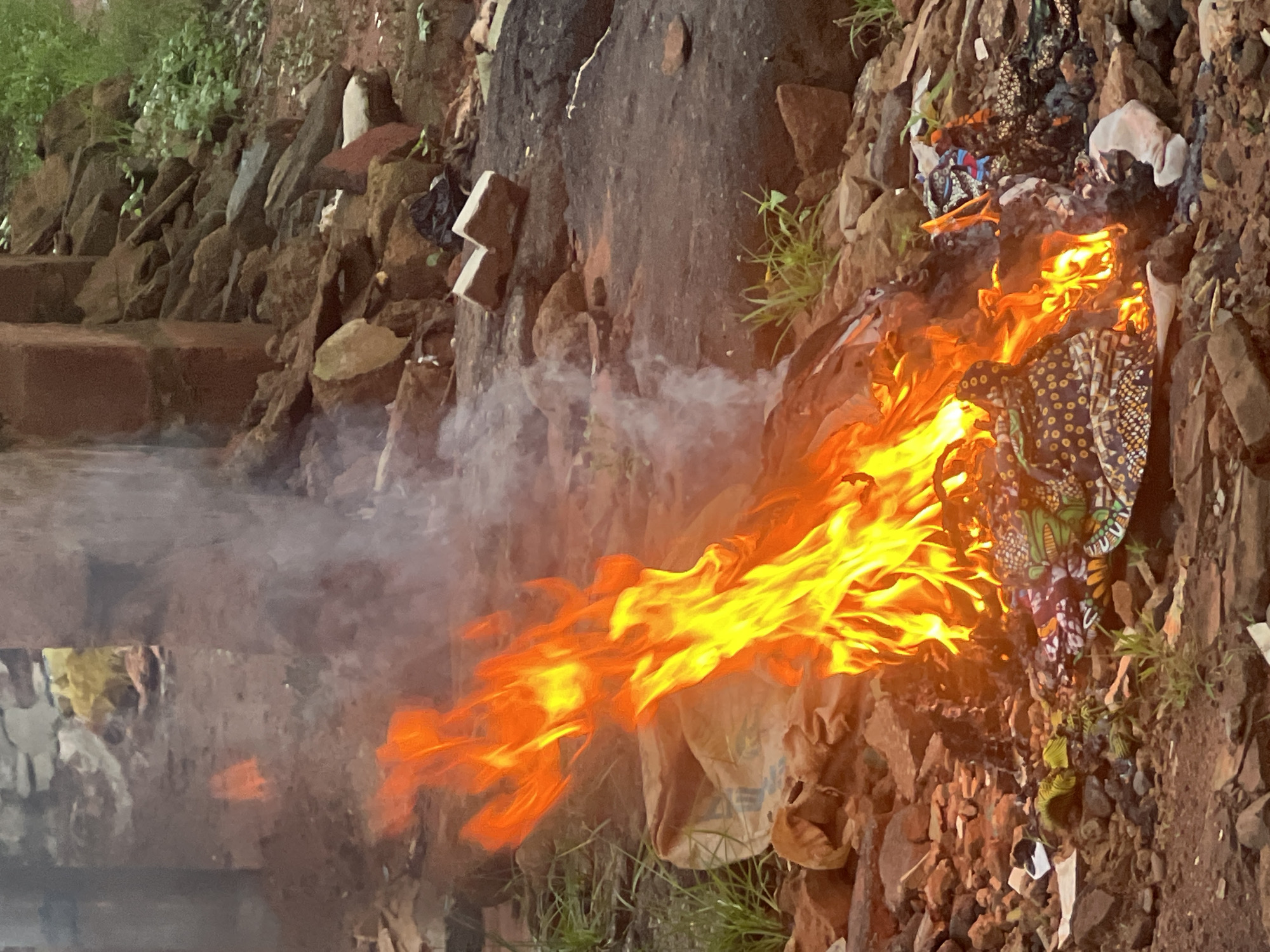 In Nkpor, Nigeria, an outdoor scene depicts a fire consuming a pile of discarded materials. The fire, with its bright orange and yellow flames, is the focal point, producing wisps of white smoke that rise into the air. Amongst the burning items are colorful fabrics, suggesting clothing or textiles, along with what appear to be plastic bags and other refuse. The ground is rough and uneven, composed of dirt, rocks, and debris, consistent with an undeveloped or neglected area within the city. In the background, there are remnants of structures, possibly unfinished buildings or ruins, made of brick and stone. The surrounding environment is sparsely vegetated, with patches of green grass and small plants struggling to grow in the harsh conditions. The overall impression is one of uncontrolled burning, likely a common method of waste disposal in the area.