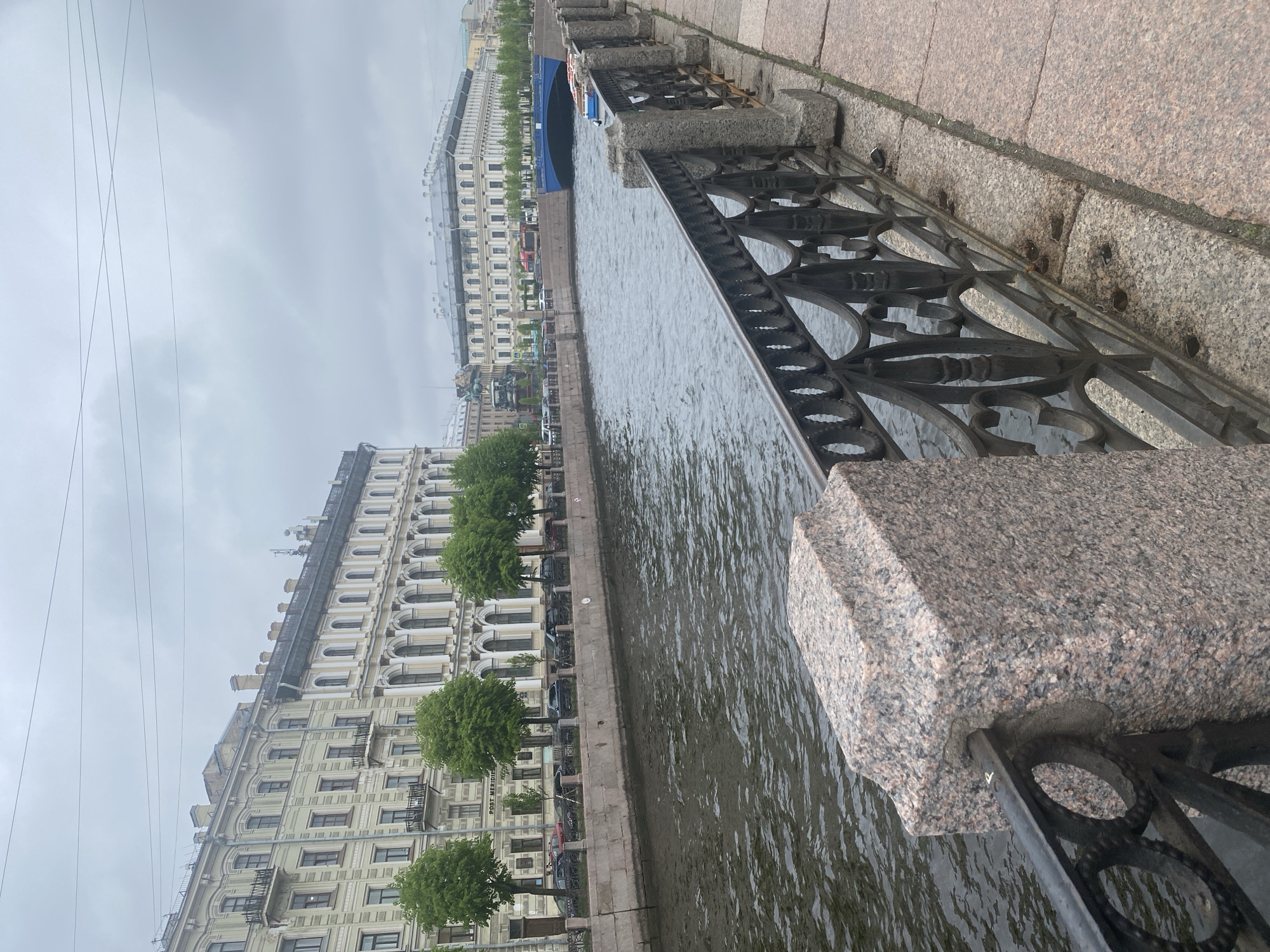 The image captures a canal view in Saint Petersburg, Russia, characterized by a cloudy, overcast sky. The foreground features a granite embankment with a decorative cast-iron fence running along the canal. The water surface is rippled, reflecting the gray sky. Across the canal, a series of multi-story buildings line the opposite bank. Trees are sparsely planted along the embankment. Parked cars are visible in front of the buildings. Power lines are strung in the sky above the buildings.