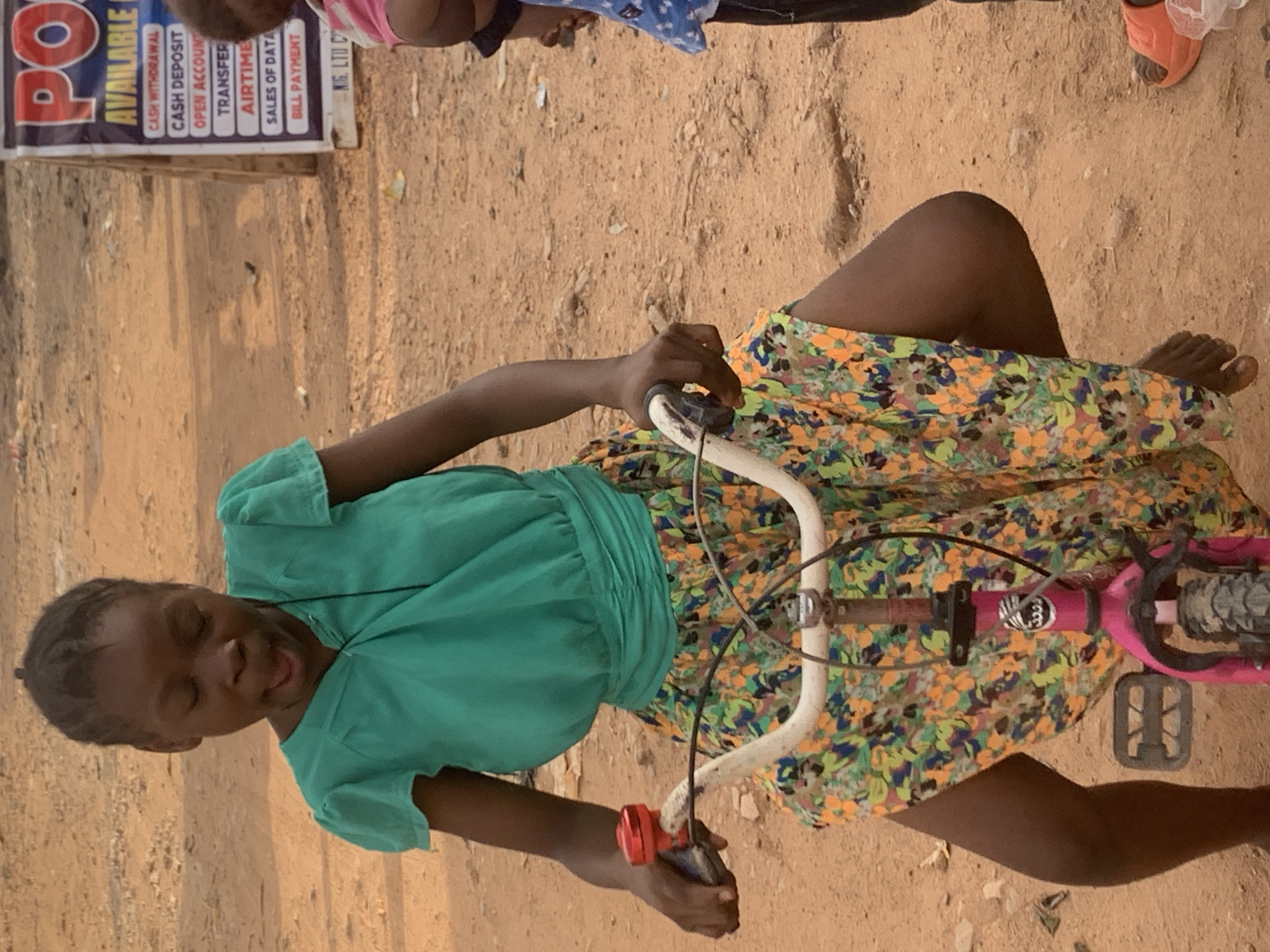 A young girl is sitting on a pink bicycle on a dirt or sandy ground in Sabon Tasha, Nigeria. The girl is wearing a green top and a colorful, patterned skirt. She is holding the handlebars of the bicycle. A sign is partially visible in the background, advertising services like "CASH WITHDRAWAL," "CASH DEPOSIT," "OPEN ACCOUNT," "TRANSFER," "AIRTIME," "SALES OF DATA," and "BILL PAYMENT." Another person is standing by the sign, but their face is not completely visible. The setting appears to be outdoors, possibly near a local business offering financial services.