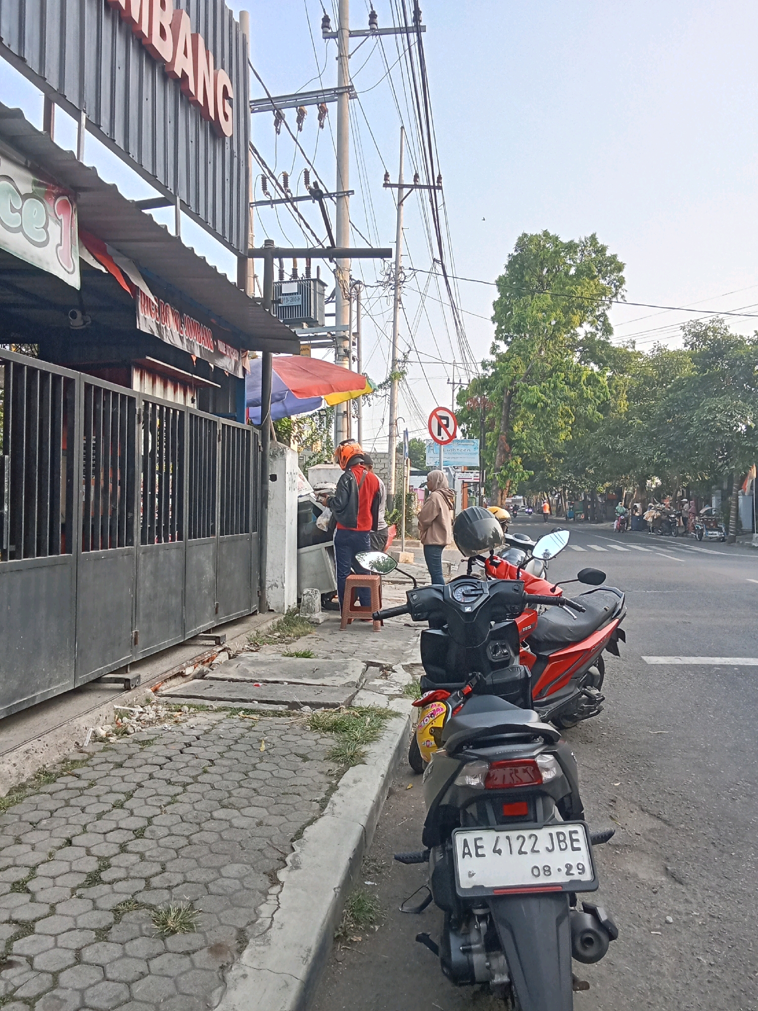 In Jombang, Indonesia, a street scene unfolds with several motorbikes parked along a sidewalk. A dark-colored motorbike with license plate "AE 4122 JBE" is prominently featured in the foreground, facing away from the camera. Behind it, a red motorbike is partially visible. The sidewalk is paved with hexagonal concrete blocks and shows signs of wear and tear, with grass growing in the cracks. Adjacent to the sidewalk is a street with ongoing traffic, indicated by the presence of various vehicles and trees lining the road. A building with a gray corrugated metal facade and the partial text "BANG" is visible on the left side of the image. Two individuals are standing near an open gate of the building, one wearing a red and black jacket with an orange helmet, and the other in a beige hijab and jacket. Electrical wires and poles crisscross the sky above the scene, and a transformer is mounted on a pole. A red and yellow umbrella is positioned near the gate. The overall impression is a busy urban environment in Jombang.