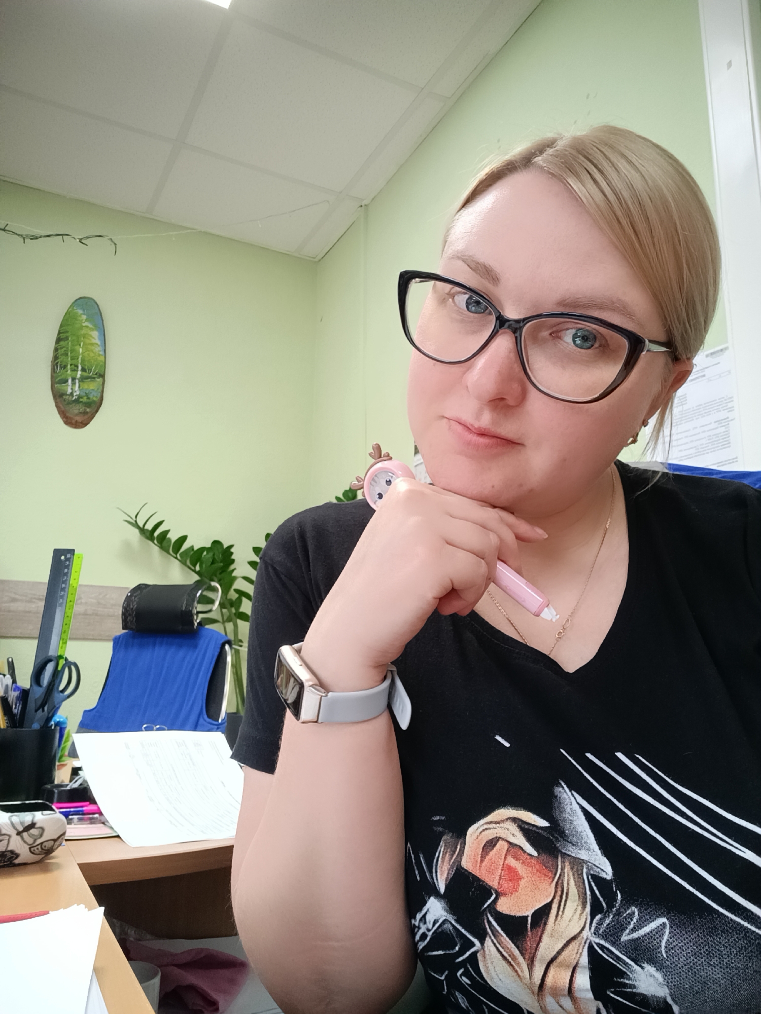 A woman is posing for a selfie in an office setting. She has blonde hair and is wearing black-framed glasses and a black V-neck t-shirt with a graphic design. She is holding a pink pen to her chin, wearing a smartwatch, and has a gold necklace on. The office has light green walls. On the wall, there is a painting of trees. A desk and chair are visible with various office supplies and a plant. A framed document is visible on the wall as well. The location is presumed to be an office in Chaikovsky, Russia.